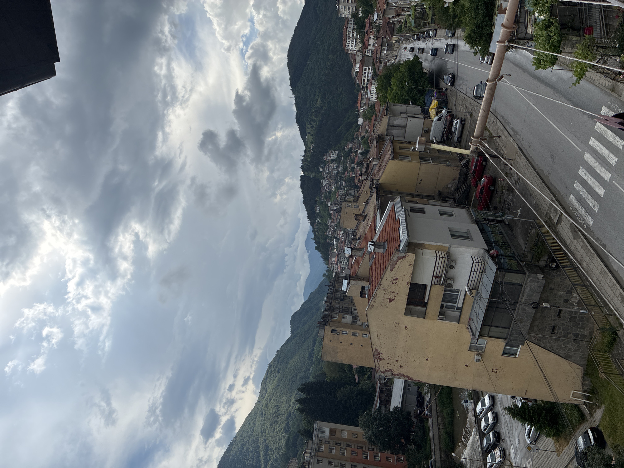 This detailed description pertains to an overhead view of a mountainous settlement in Vlahovo, Bulgaria, observed under an overcast sky. The image captures an urban environment nestled within a valley, with buildings extending up verdant, tree-covered hillsides.
The sky is predominantly grey with dense cumulus and stratus clouds, indicating either recent precipitation or an impending weather event. Patches of brighter sky are visible where the cloud cover is thinner.
In the lower left quadrant, a small, dark-colored river or stream flows, featuring a visible weir or small waterfall. Adjacent to the river is a paved area with several parked vehicles, including multiple light-colored cars and a dark SUV. Further into the left, a multi-story apartment building, partially visible, exhibits an orange-red and off-white facade. Next to it, a distinct multi-story building with a weathered, peeling yellow exterior stands on a slope, supported by a stone foundation at its base. This building features multiple windows, balconies, and an enclosed glass-fronted extension on a lower level.
A central asphalt road, visibly wet and reflective, traverses the scene from the lower center-right, curving towards the upper left. Numerous vehicles, both parked and in motion, are present on this road. A clearly marked pedestrian crossing (zebra stripes) is painted on the road in the lower foreground. Overhead, a thick, light-colored pipe or beam spans horizontally across the street, supported by wires or ropes, possibly part of utility infrastructure.
Residential and commercial buildings, predominantly multi-story structures with orange or red-tiled roofs, line both sides of the main street and are densely clustered on the lower slopes of the surrounding hills. Many buildings display signs of age, including weathered facades and peeling paint. Trees and lush green foliage are interspersed throughout the urban landscape, particularly along the riverbank and on the higher slopes.
The background is dominated by imposing, heavily forested mountains on both the left and right sides. On the right-hand mountain, numerous houses are visible, built into the ascending terrain. Distant mountains with hazy outlines are discernible further in the background, beneath the cloudy horizon. No individuals are clearly identifiable or engaged in specific actions within the visible street or building areas.