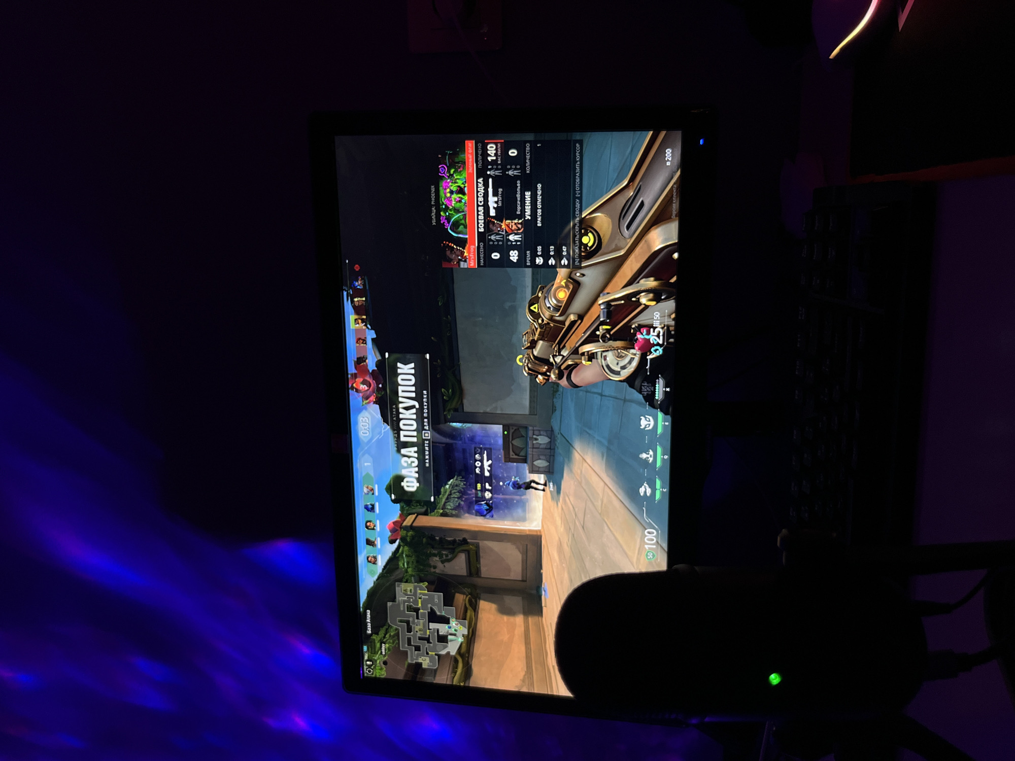 The image shows a computer screen displaying the game Valorant. The HUD elements indicate it is the "buy phase" of a round. The player's health is at 50/100, and their shield is full at 100. The screen also displays kill feed information, with the player character being named "MrsFrog". The game interface is displayed in Russian. A microphone is partially visible in the foreground. A keyboard is also visible to the right of the screen. Blue light illuminates the left side of the wall, providing a light to the darkness of the image. There is no explicit indication of location in the city of Rostov-on-Don, Russia.
