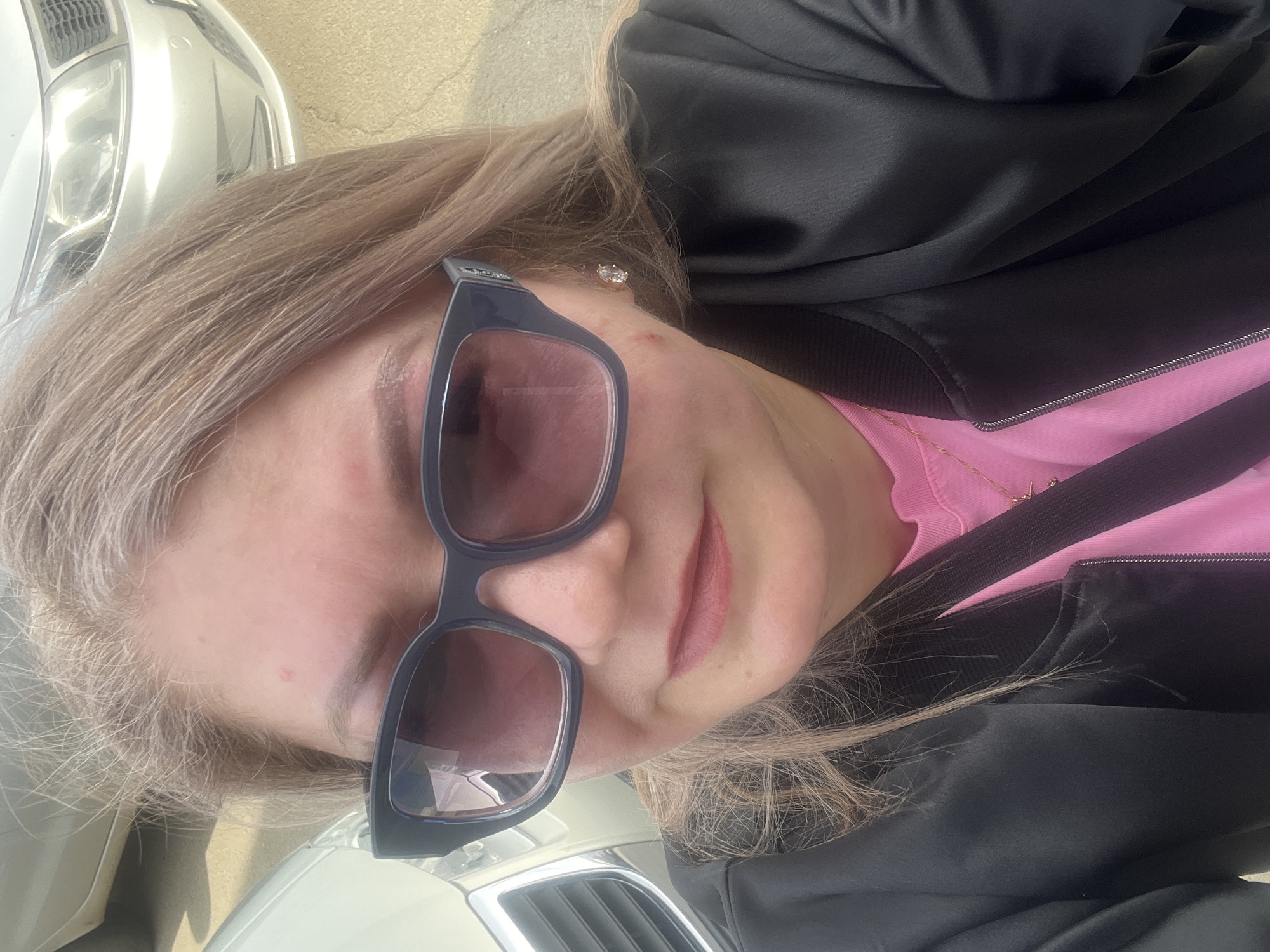 A close-up, rotated portrait shows a person, likely female, from the chest up, positioned against the backdrop of a white or silver car. The person has light, possibly blonde or light brown hair, which appears slightly disheveled. Large, dark-framed sunglasses with reddish-purple tinted lenses cover a significant portion of the face, obscuring the eyes. The person has natural-toned lips.
They are wearing a bright pink top, visible under a black bomber jacket or similar outerwear with ribbed cuffs and a zipper. A thin gold chain with a small, delicate pendant is visible around the neck. A single silver stud earring is seen on the earlobe.
The background primarily features parts of a car, including a headlight and grille, suggesting an outdoor setting, likely a parking area or street. The lighting indicates it is daytime and sunny. No specific activity beyond posing for the photograph is evident. There is no visible text in the image. The location context is Izhevsk, Russia.