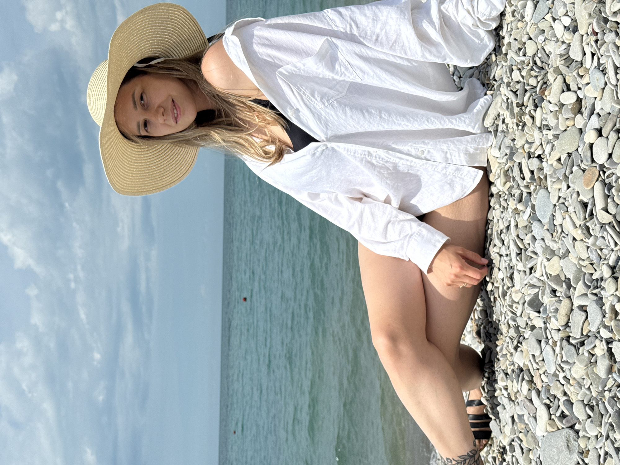 A young woman wearing a white oversized shirt and a large straw hat is sitting on a pebble beach. She is wearing a black swimsuit underneath the shirt, and has a tattoo on her left ankle. Her legs are crossed, and she is looking towards the camera with a slight smile. The sea is visible behind her, with a cloudy sky overhead. The location is the pebble beach in Nebug, Russia.