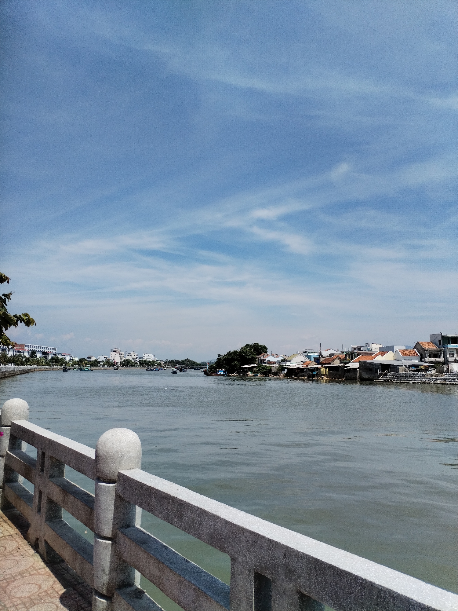 The image captures a wide, calm river in Nha Trang Ward, Vietnam, under a partly cloudy blue sky. In the foreground, a grey concrete railing with rounded posts runs along a tiled walkway. The railing has rectangular openings and a speckled texture. The river occupies the middle ground, with numerous small boats scattered across its surface. On the far bank, a town is visible, featuring a mix of residential buildings and some larger structures. The buildings display a variety of architectural styles and roof types, with some appearing to be older, low-lying structures and others more modern, multi-story buildings. Trees and vegetation are interspersed with the buildings. The overall impression is of a serene, sunny day in a riverside community.