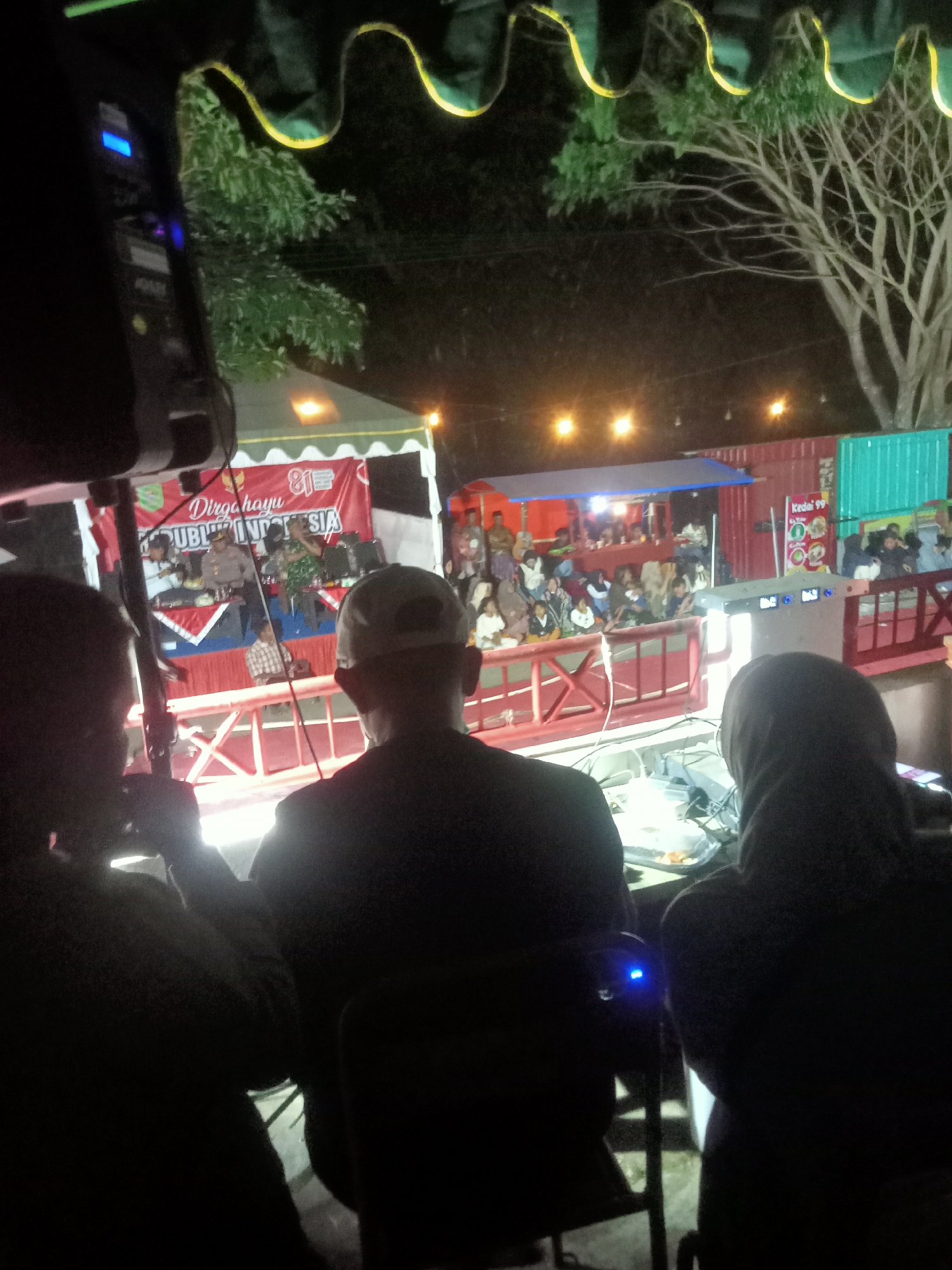 A night-time outdoor event is underway. In the foreground, three individuals are seated facing away from the camera. The person on the left is partially obscured, while the person in the center wears a baseball cap and the person on the right is wearing a head covering. They are positioned behind a table with electronic equipment. In the middle ground, a stage area is set up with a red and white banner displaying "Dirgahayu REPUBLIK INDONESIA" and "81". Several people are on the stage, including one in a police uniform and others in casual attire. A crowd is gathered in front of the stage, seated or standing. The background shows a line of food stalls or vendors, one with a sign that reads "Kedai 99". Trees and a dimly lit, possibly barren, tree are visible in the upper portion of the image. The overall lighting suggests artificial illumination from the stage and stalls. The scene appears to be in a public space in Pule, Indonesia.