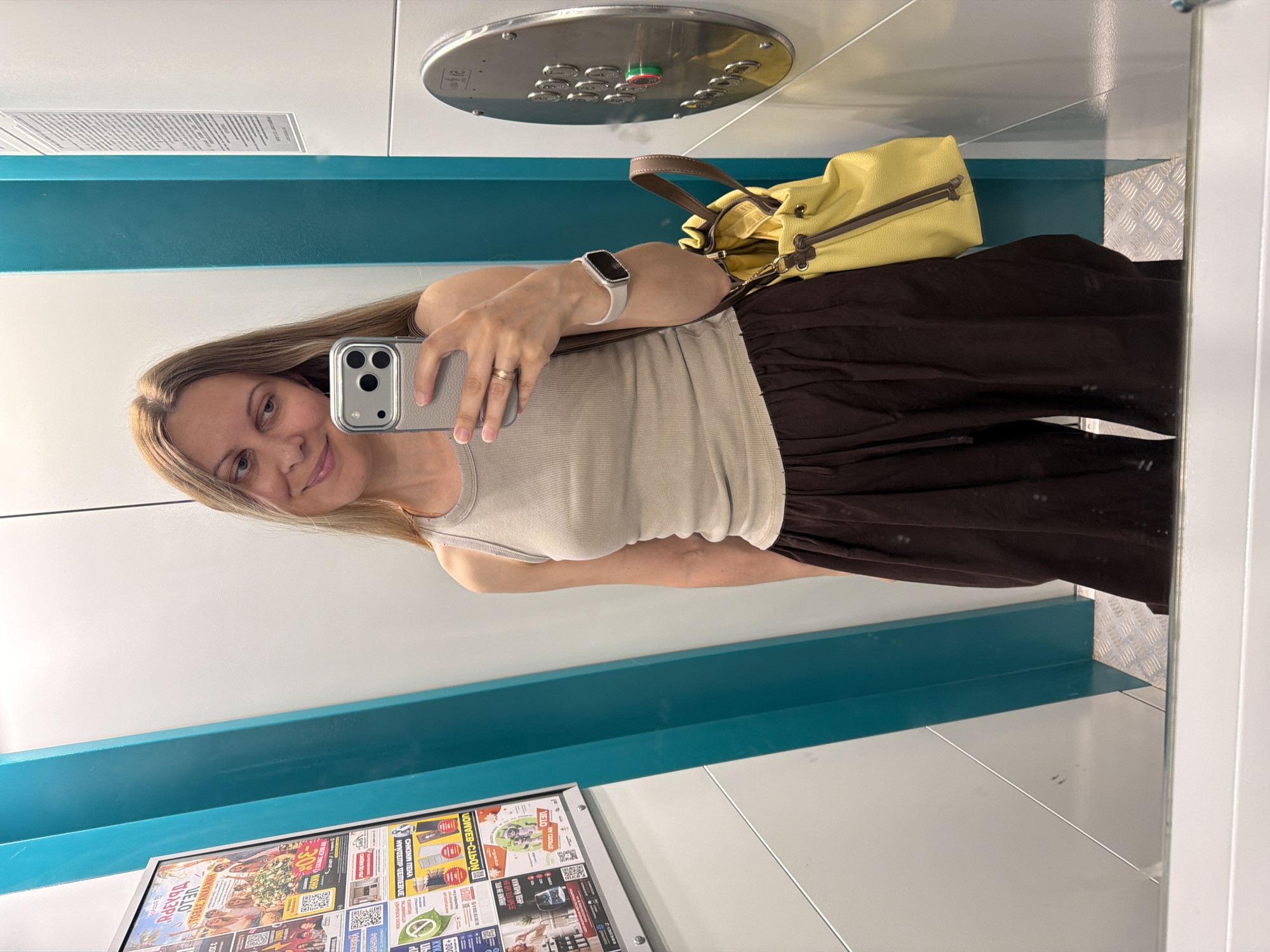 The image shows a young Caucasian woman with long blonde hair taking a mirror selfie. She is wearing a light brown ribbed tank top and dark brown wide-leg pants. She has a light grey phone in her right hand, and a yellow drawstring bag with brown straps is slung over her left shoulder. She is also wearing a smartwatch on her left wrist. The background appears to be the interior of an elevator, with teal and white walls and a metallic panel with elevator buttons visible on the right. A poster with text and images is mounted on the left wall. The flooring is a metallic diamond plate pattern. The overall scene suggests the woman is in a public space, possibly an apartment building or office complex, in the city of Izhevsk, Russia, indicated by the text on the poster which appears to be in Cyrillic script.