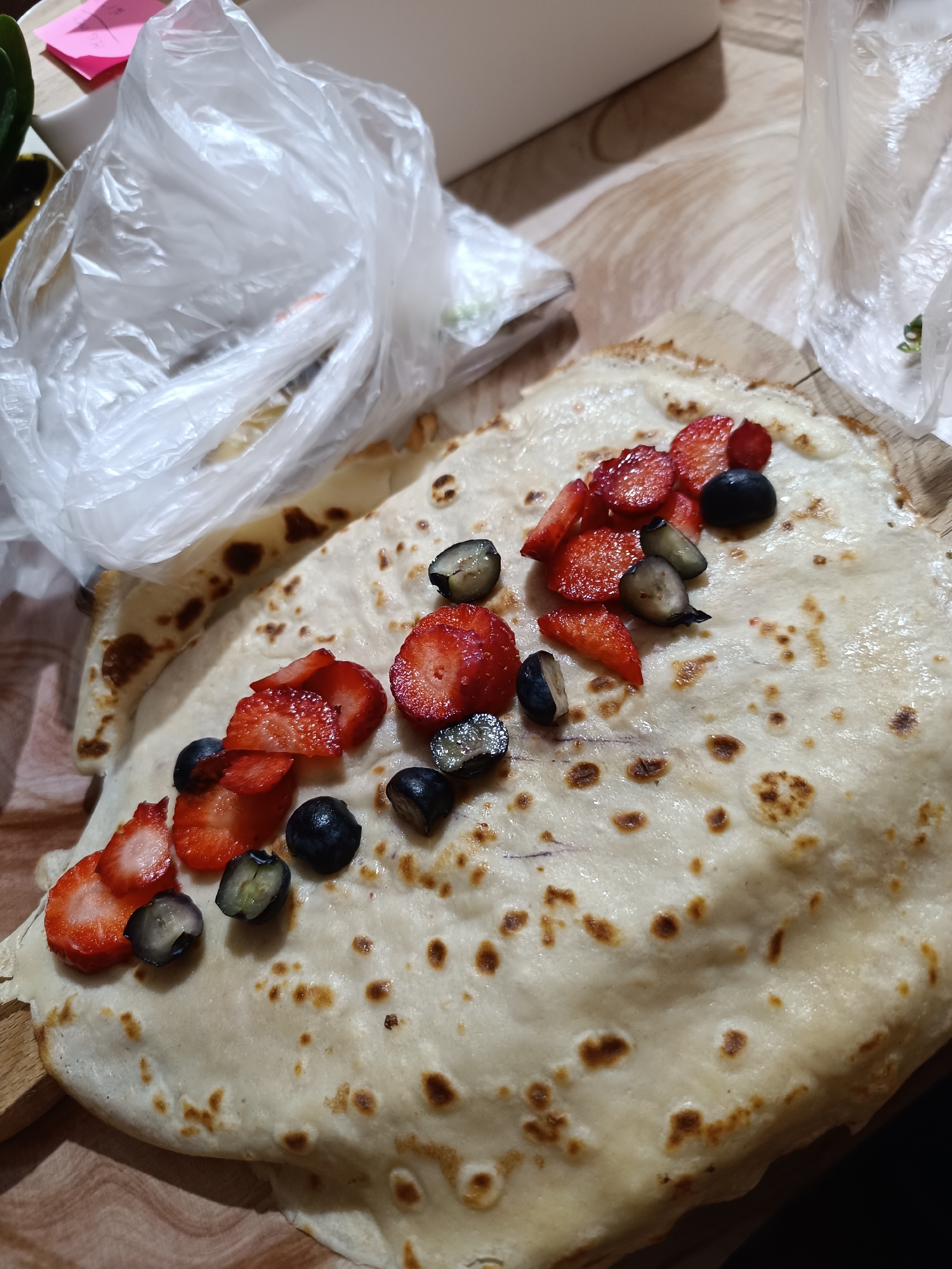On a wooden surface, a large, thin pancake or crepe is topped with sliced strawberries and blueberries. The pancake is light in color with browned spots. The strawberries are bright red, and the blueberries are dark blue/purple. To the left and right of the pancake are plastic bags, one of which is partially obscuring a small plant. The location is not evident in this image. The image does not contain any information that suggests the scene is located in Pleven, Bulgaria.