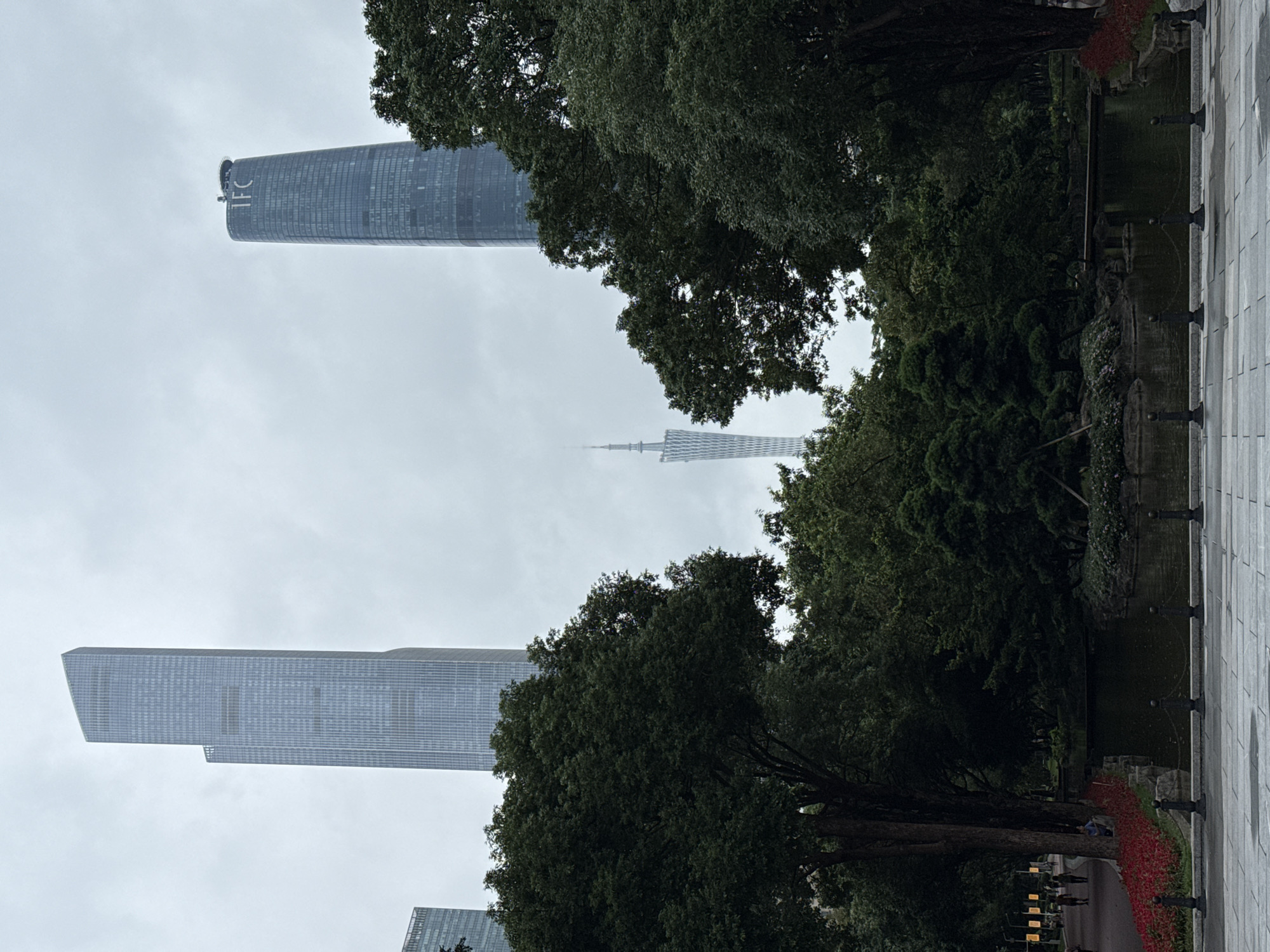 The photo depicts a view in the Tianhe District of Guangzhou, China. In the background, two skyscrapers, the Guangzhou CTF Finance Centre and the Canton Tower, are visible through trees on an overcast day. In the foreground, there is a pond surrounded by landscaping. A stone path runs along the side of the pond.