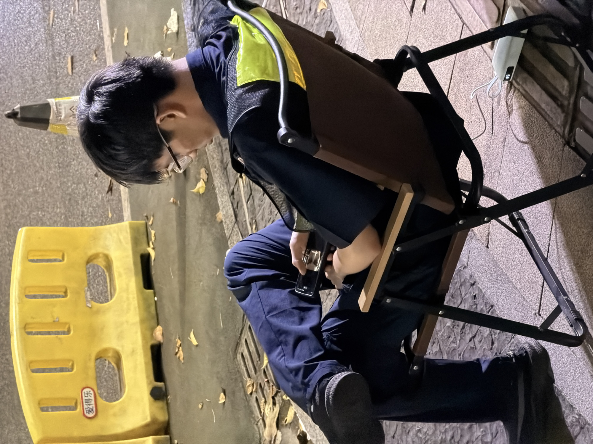 A person wearing a dark blue uniform with a reflective yellow vest is seated on a portable folding chair outdoors, looking down at a handheld device. The individual's legs are crossed, and they are wearing dark trousers and black shoes. The background shows a yellow barrier with the Chinese characters "爱得乐" visible. There are fallen leaves scattered on the ground, suggesting an outdoor setting, possibly a street or public area in Chancheng District, China. A black traffic cone is visible in the distance. A white electronic device with a wire is placed on the ground near the chair. The lighting suggests it is evening or night.