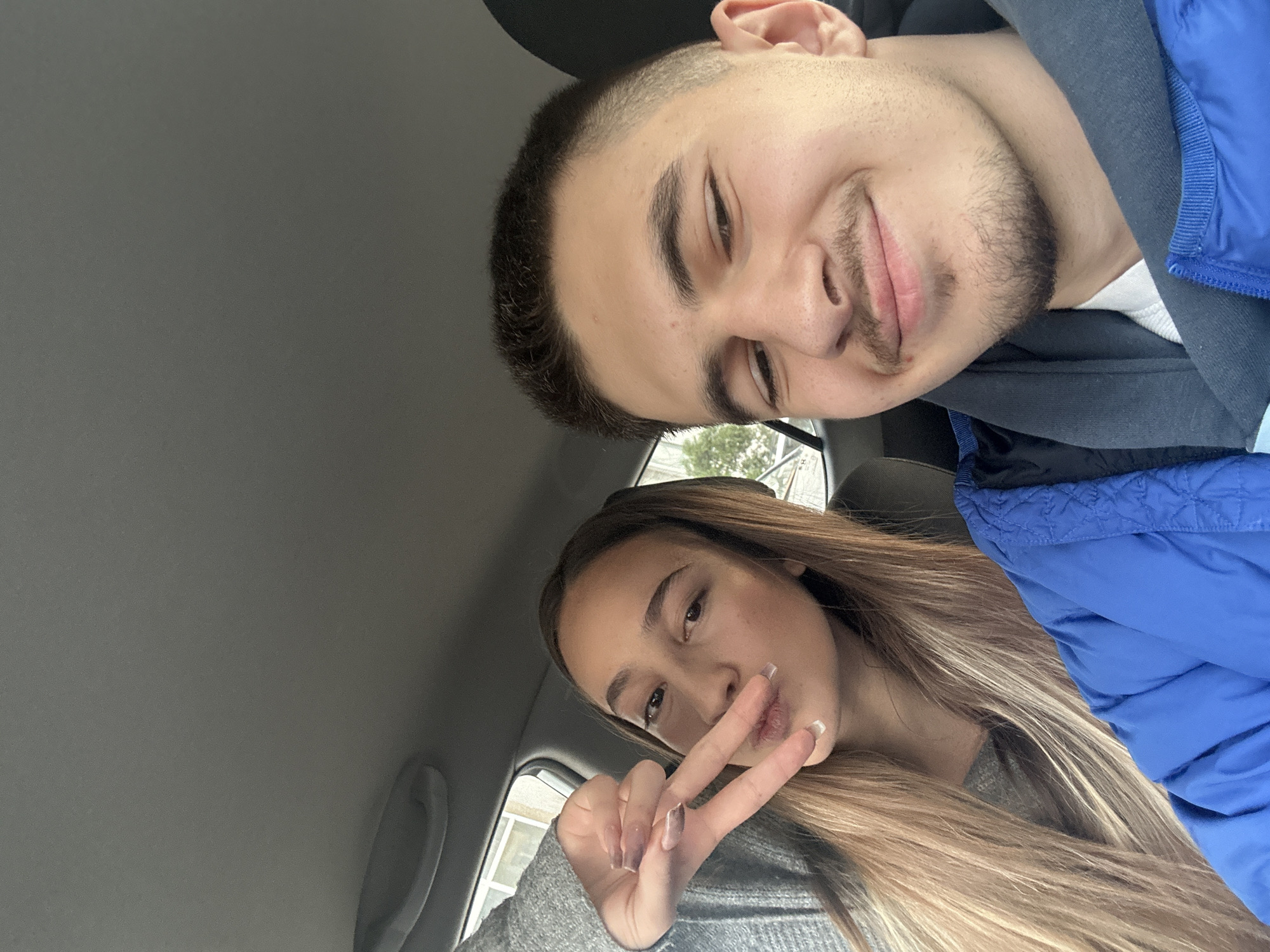 The image depicts two people, a male and a female, inside a car. The male is positioned at the top of the frame, closer to the car's roof. He has short dark hair, a light beard, and is wearing a blue jacket over a grey hooded sweatshirt and a white t-shirt. The female is positioned below him. She has long blonde hair and is wearing a grey sweater. She is making a peace sign with her fingers. The interior of the car is visible. There is no information to support that the location is Sofia, Bulgaria. The scene is well-lit.