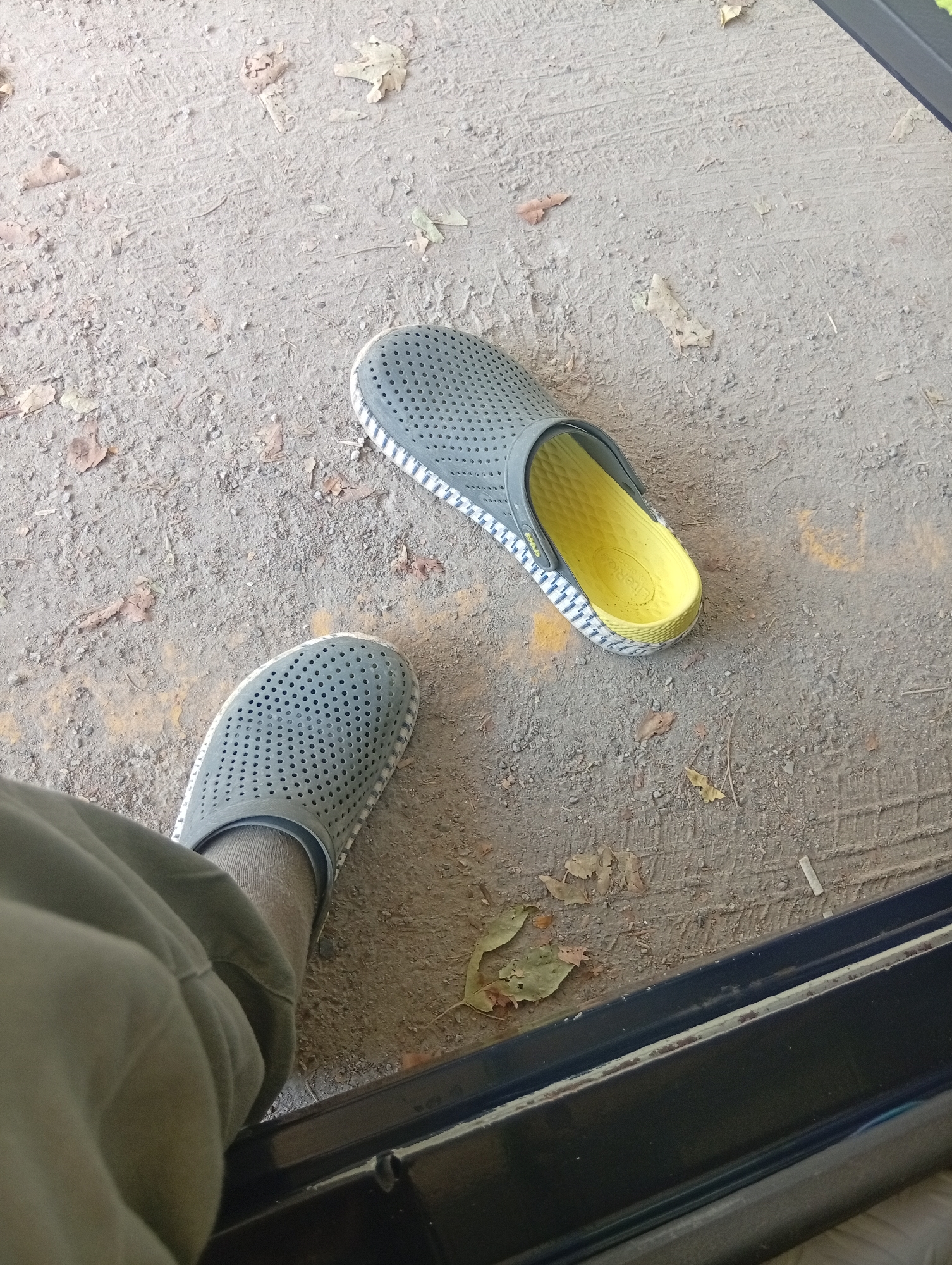 A pair of grey perforated clogs with white and blue striped soles and bright yellow insoles are situated on a dusty, outdoor surface. One clog is closer to the camera, partially obscured by the leg of a person wearing khaki pants and a grey sock. The other clog is further away, facing in the opposite direction. The ground is a mixture of dirt, gravel, and scattered fallen leaves. The scene appears to be located outdoors, possibly on a sidewalk or pathway, near a dark-colored edge that could be part of a building entrance or structure. No specific landmarks or indicators of Tashkent, Uzbekistan, are visible in the image.