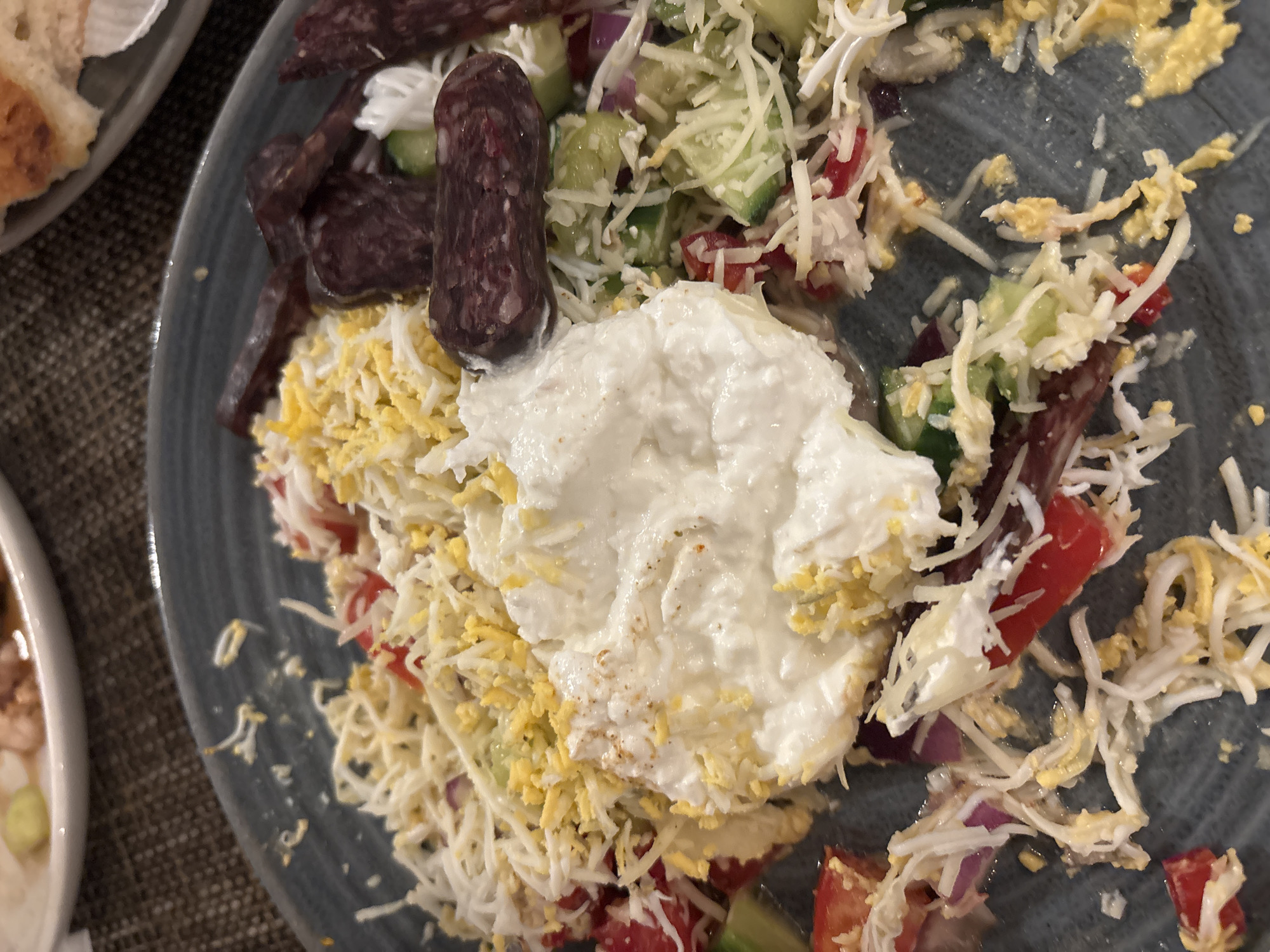 A top-down shot depicts a plate of unfinished salad. The salad includes chopped cucumbers, tomatoes, red onions, shredded cheese, shredded egg, and slices of sausage. A dollop of white cream is situated at the center of the salad. The plate is dark gray. Part of another plate containing a piece of bread is visible on the left of the frame. Based on the food presented, the image may have been taken in Sofia, Bulgaria.