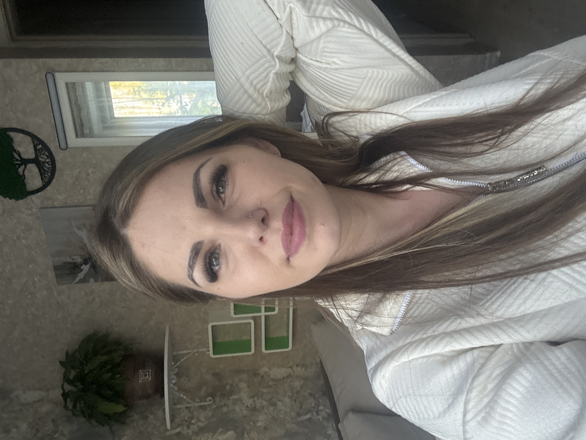 A woman with long, light brown hair is shown in a close-up shot. She is wearing a white jacket with a textured pattern. Her eyes are green, and she has visible eyelashes and eyebrows. She has a nose piercing. In the background, there is a window and a decorative tree. A potted plant is also visible. There is nothing to indicate the location is Раздолье, Russia.