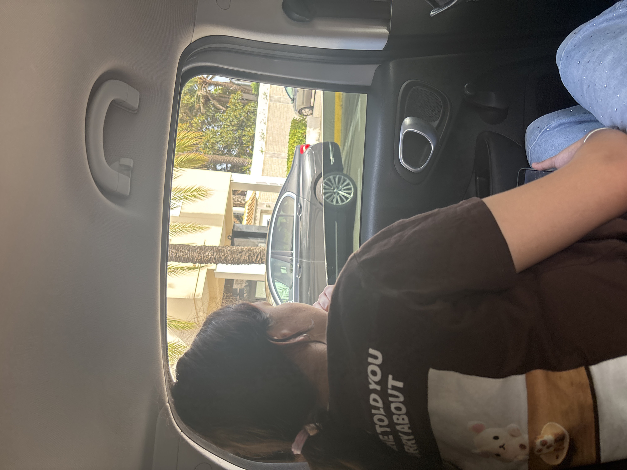 The image shows a person sitting in the backseat of a car, looking out the window. The person has long, dark hair tied back with a pink scrunchie and is wearing a dark brown t-shirt with white lettering that reads "ME TOLD YOU RRY ABOUT". A small stuffed animal and a miniature cake are visible on the t-shirt. Through the car window, a dark gray sedan is visible driving on a road, with buildings and palm trees in the background. The scene appears to be in a sunny, urban environment. The location is Urbanización Cabo Roig, Spain.