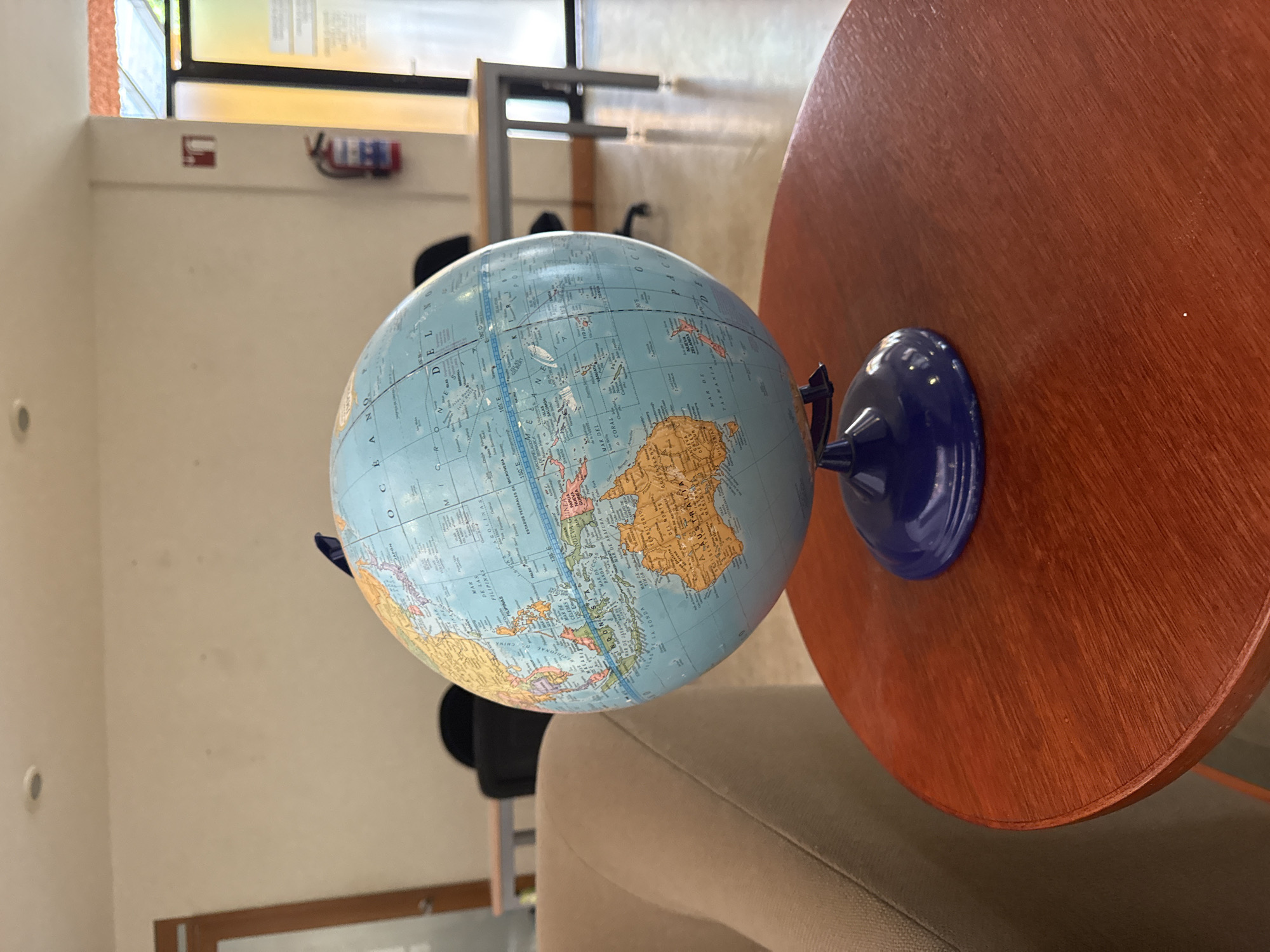 The image shows a globe resting on a round, wooden table. The globe is light blue with visible continents and oceans. The base of the globe is dark blue. The table is a reddish-brown color with a smooth surface. The background is blurred, showing a room with white walls, a window, and chairs. A fire extinguisher is visible on the wall in the background. The setting appears to be indoors, likely in a cafe, home, or office in San Andrés Cholula, Mexico.