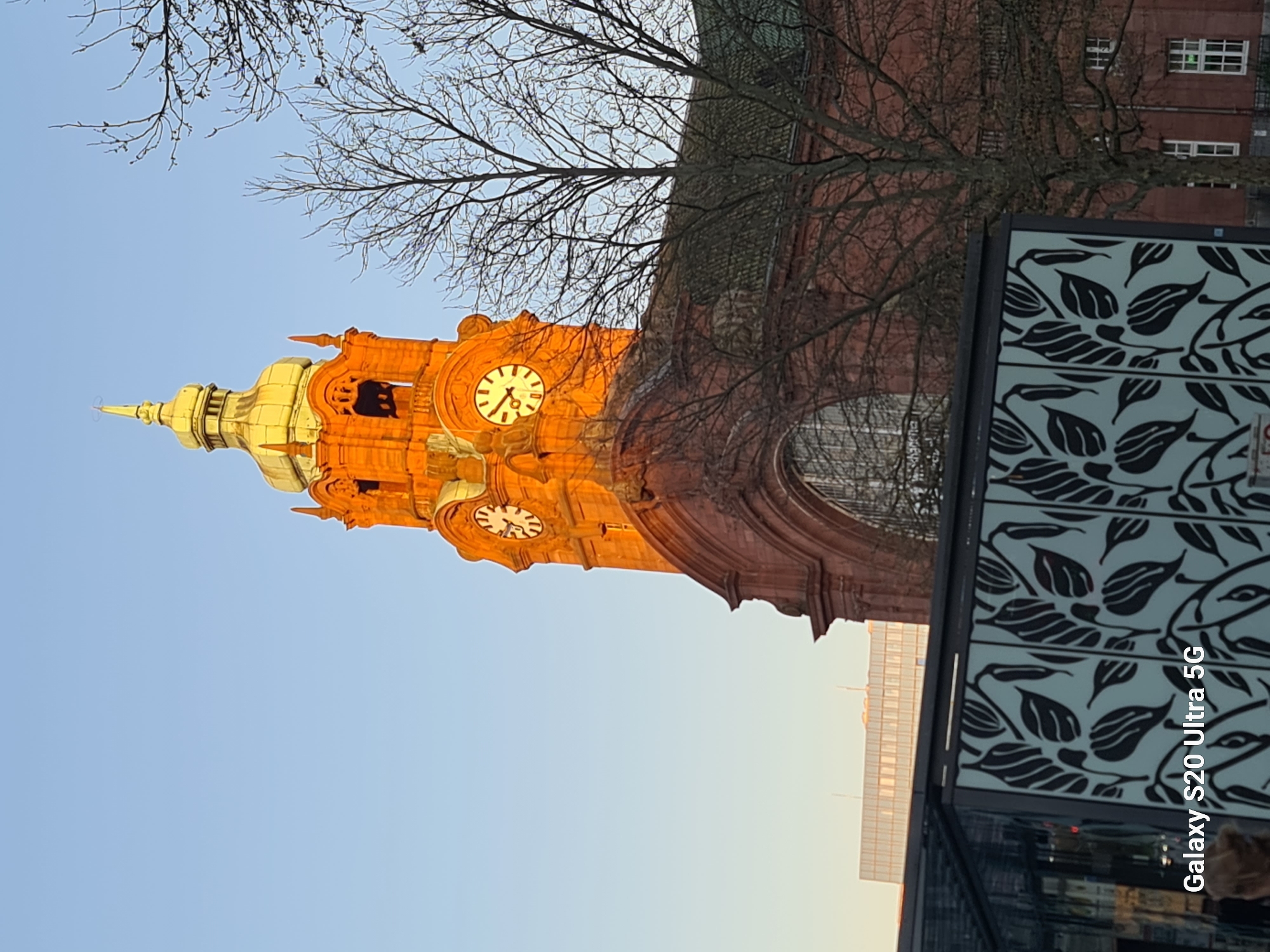 The image shows the Marktkirche (Market Church) in Wiesbaden, Germany, from a low angle. The church's orange brick facade is illuminated by the setting sun, contrasting against the light blue sky. Bare tree branches are visible in the foreground, partially obscuring the view of the church. A modern structure with a leaf-patterned design is present in the lower right corner. Another building is partially visible in the background. Two clocks are mounted on the tower. The text "Galaxy S20 Ultra 5G" is in the lower right corner, indicating the device used to capture the image.