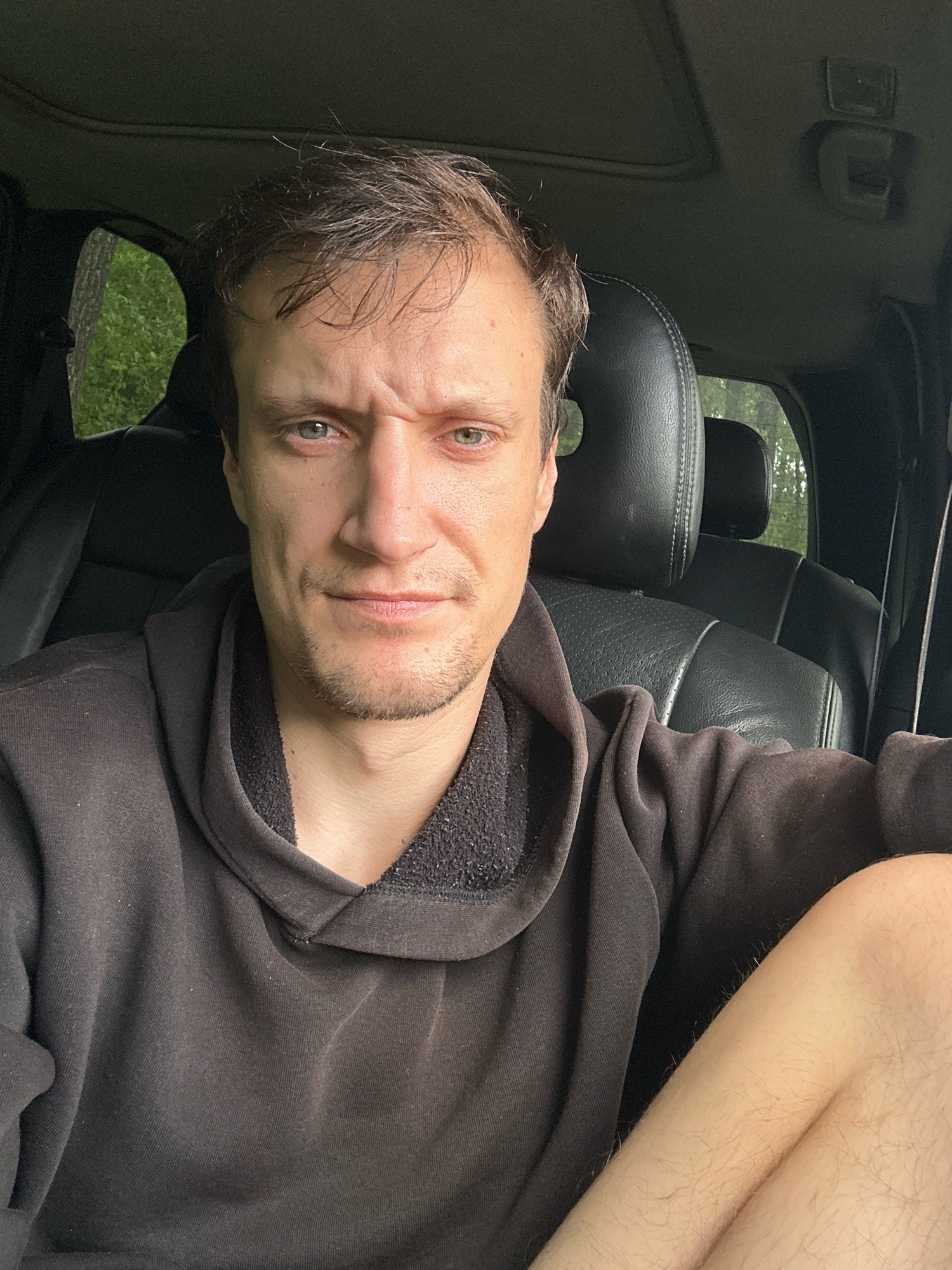 The image is a selfie taken from inside a vehicle, likely an SUV, with a dark interior. The subject is a Caucasian male with short brown hair and a light beard, looking directly at the camera with a furrowed brow and an intense expression. He is wearing a dark, hooded sweatshirt. The background visible through the windows suggests a natural environment, possibly a forest or wooded area, with green foliage. The interior of the car features black leather seats and a dark headliner. There are no discernable elements that would specifically place the location in Makiv, Ukraine.