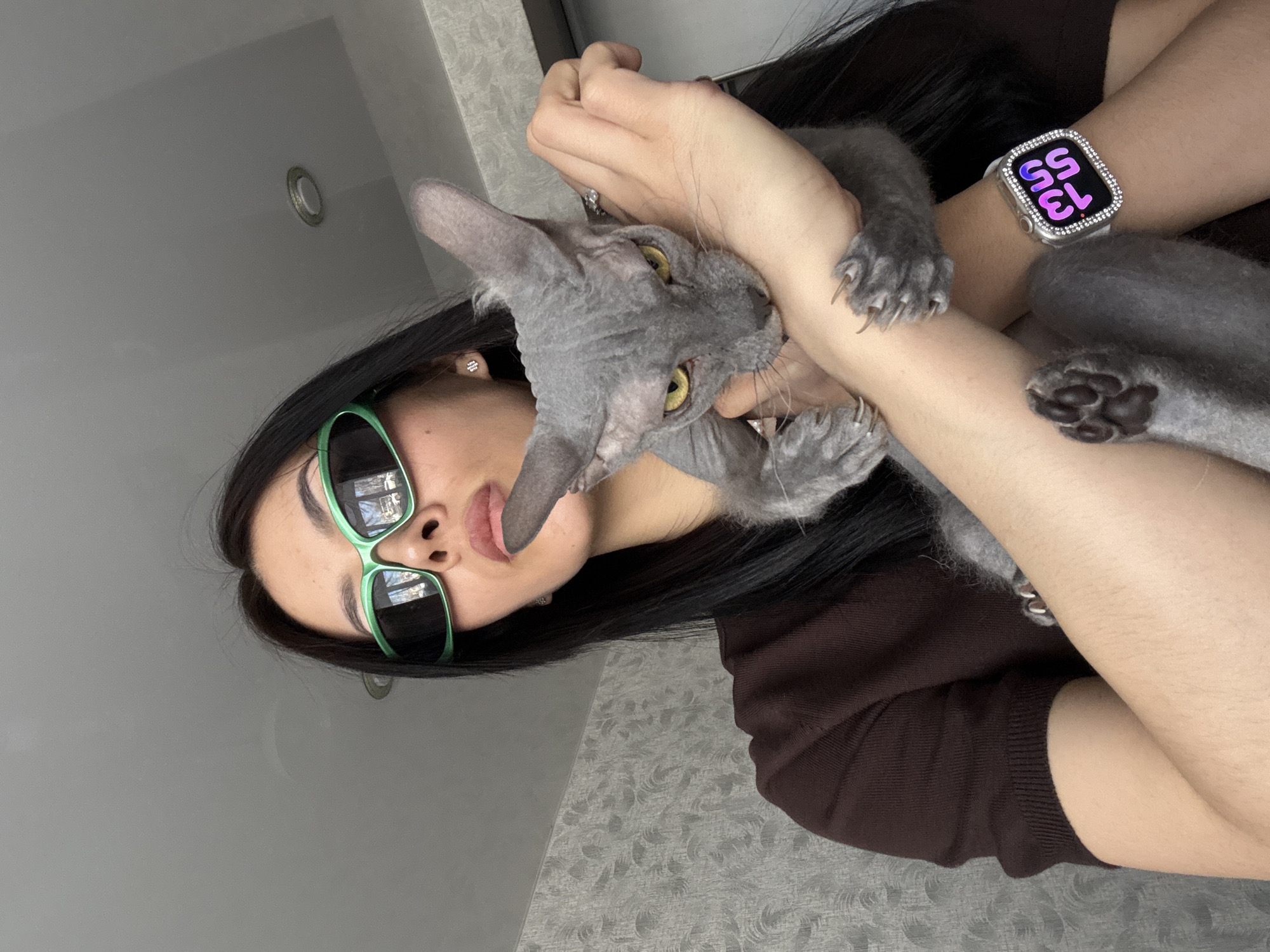 A woman with long, dark hair and green-framed sunglasses is holding a gray, hairless cat. The woman wears a dark brown sweater and a silver smartwatch with a sparkly bezel. The cat has yellow eyes and is clinging to the woman's arms. The background is a neutral gray wall. The location cannot be determined to be Votkinsk, Russia based on the image.