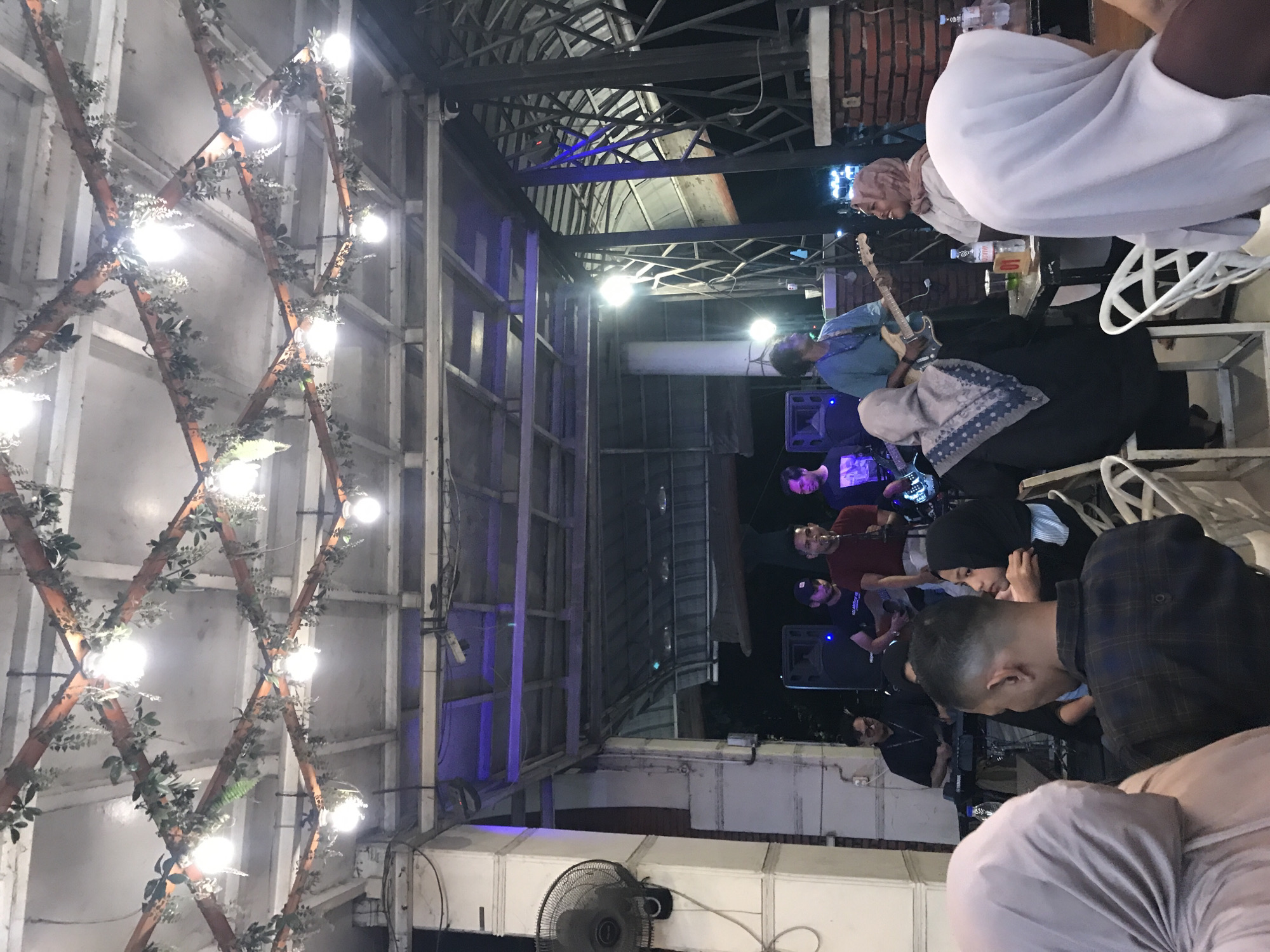 The image depicts a scene at an outdoor venue in Lhokseumawe, Indonesia. A band is performing on a stage or platform. The band appears to have a guitarist, bassist, and drummer, among others.
Several people are in attendance, seated at tables. Some are facing the performance area. The individuals include those wearing hijabs.
The venue has metal scaffolding and decorated with lights. There is a fan.