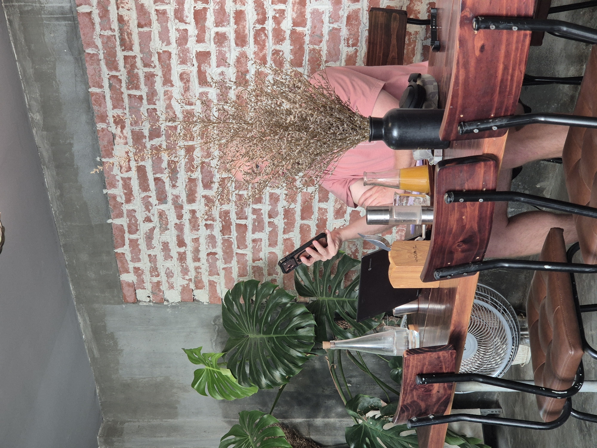 A person wearing a pink shirt is sitting at a wooden table in a cafe in Da Nang, Vietnam, holding a smartphone. The table is laden with items including a laptop, a glass bottle of water, a small wooden box, two glasses containing liquids, a black vase with dried flowers, and a pair of headphones. The cafe's interior features a textured concrete wall on the left and a red brick wall on the right, with several large green plants adding to the ambiance. Two chairs with brown leather seats and dark metal frames are positioned at the table. A fan is visible behind the chairs. The lighting suggests it is daytime.