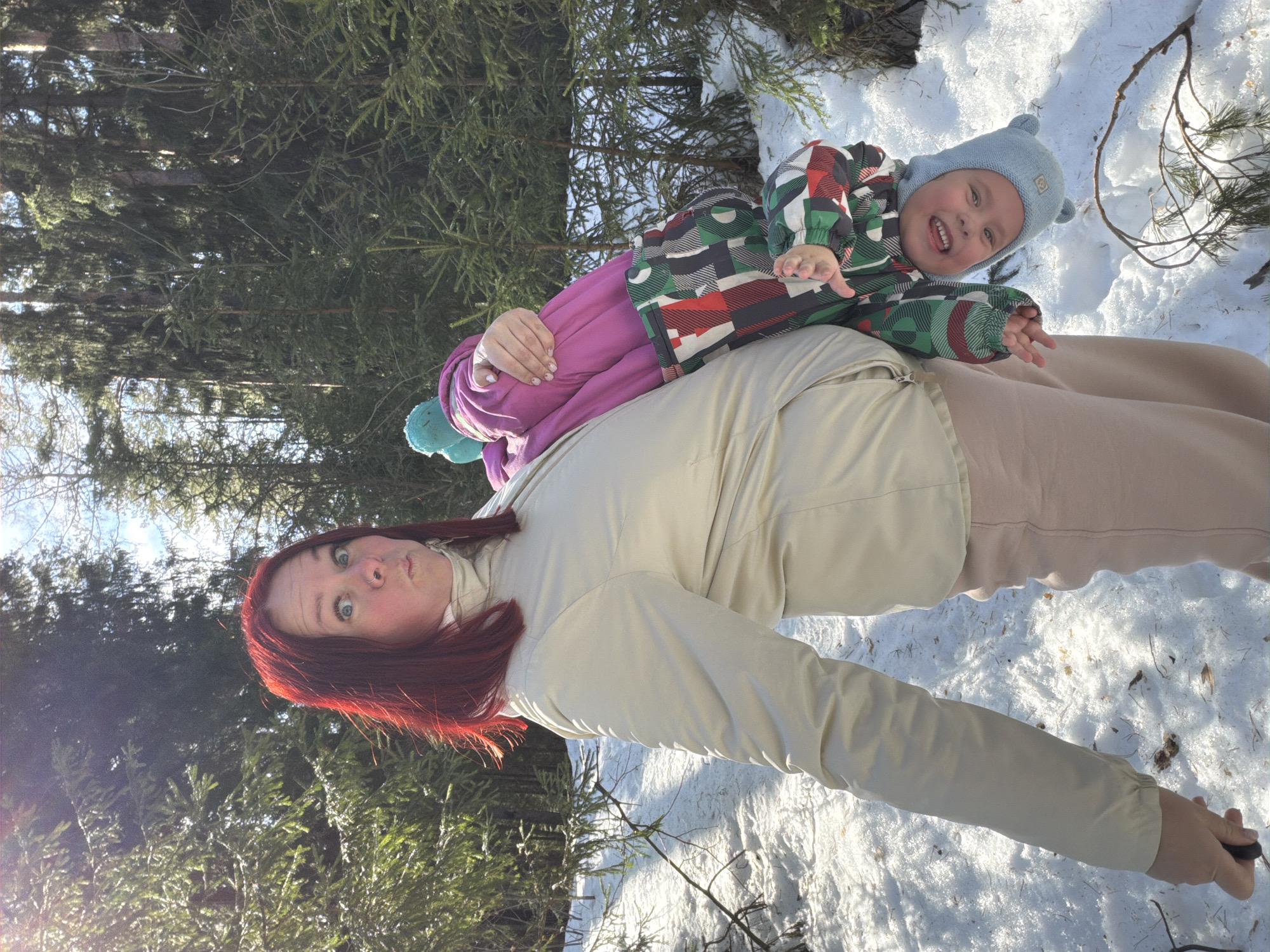 An outdoor scene in a snowy forest environment within Chaikovsky, Russia, features a woman playfully holding a child upside down. The woman, positioned centrally, has shoulder-length red hair, blue eyes, and a nose piercing, and exhibits a pursed-lip expression. She wears a light beige long-sleeved jacket with a high collar and light pink trousers. Her left hand, visible at the bottom left, holds a dark cylindrical object. The child, a toddler, is dressed in a bright pink top, a multicolored winter suit with a geometric pattern (green, red, white, dark gray), and a light blue beanie with ear-like protrusions. The child's head is inverted towards the lower right of the frame, showing a wide, open-mouthed smile with visible teeth. The child's feet rest against the woman's left hip/side. The ground is covered in white snow, reflecting bright sunlight, with visible scattered pine needles and small branches. The background consists of a dense coniferous forest with numerous evergreen trees of various sizes under a bright sky.