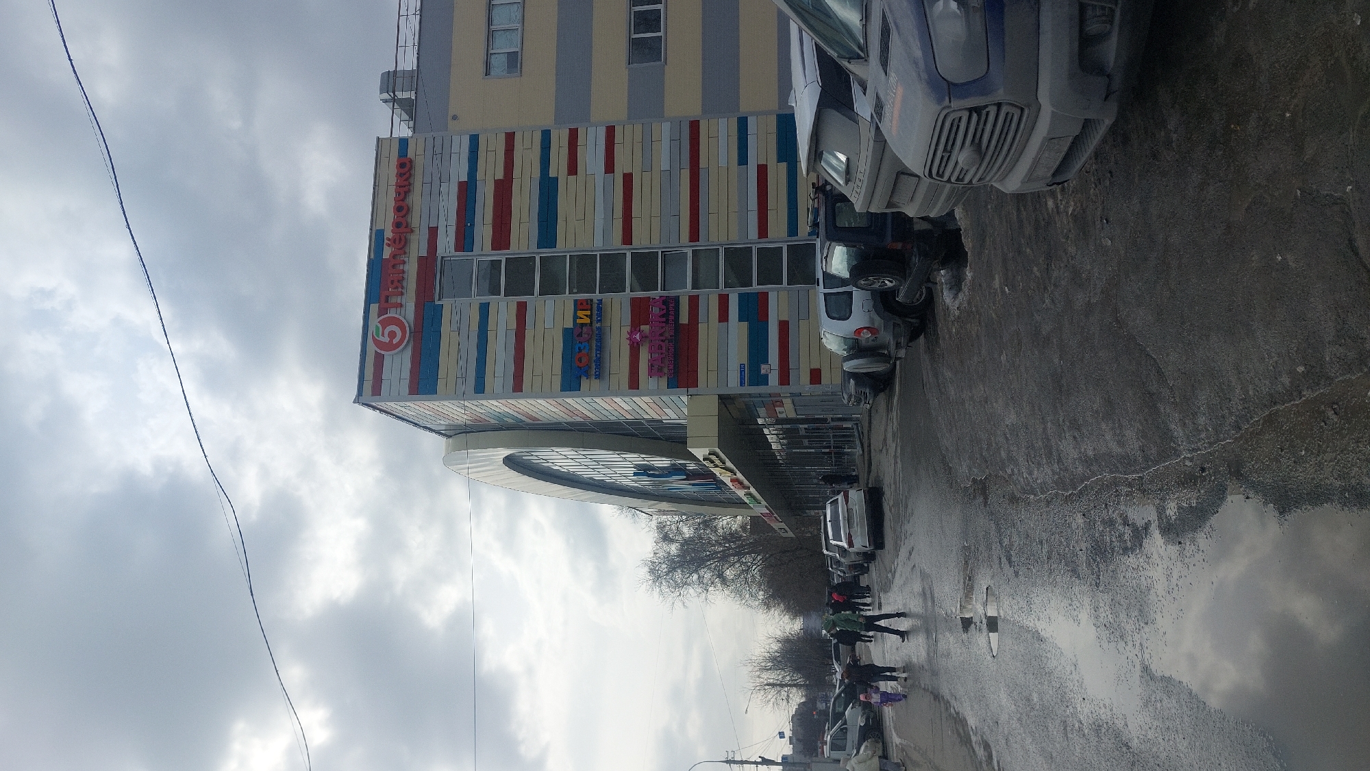 The image depicts a street scene in Novosibirsk, Russia, likely during the off-season due to the cloudy sky, bare trees, and wet pavement. A multi-story building dominates the scene, displaying a facade with alternating red, white, and blue horizontal stripes. The building's signage includes the logo of "Pyaterochka", a Russian grocery chain, and other commercial signage, indicating retail businesses. Several cars are parked along the road, and pedestrians are walking on the sidewalk, some of them are dressed in winter clothing. The street surface appears wet with puddles, reflecting the overcast sky. Utility poles and wires are visible overhead.