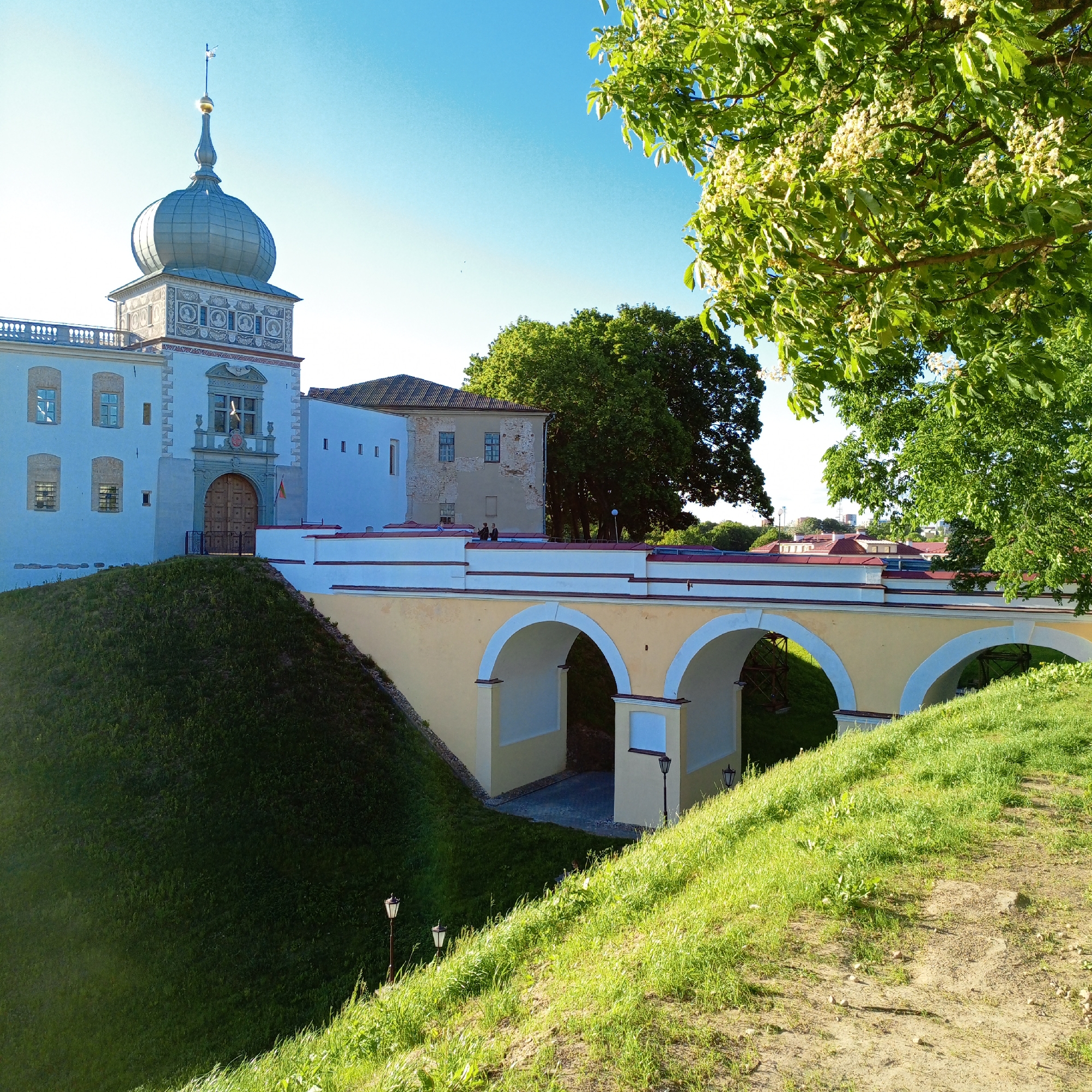 The image shows a castle or fortified structure in an urban setting. The scene is illuminated by bright sunlight, creating strong contrast. The sky is clear and blue.
In the foreground is a grassy slope leading up to a cream-colored arched bridge with a white parapet. A green tree is visible to the right, partially framing the scene.
The castle or fort features a light blue exterior with a prominent onion-shaped dome on a square tower. Details of the architecture include decorative elements on the tower, visible windows, and a grand entrance door. A smaller building is connected to the main structure, with a terracotta-colored roof.
Two figures are standing on the bridge near the center of the frame, appearing as small silhouettes against the bright backdrop.
The overall scene suggests a historical site, possibly a tourist attraction, located in a city environment.
