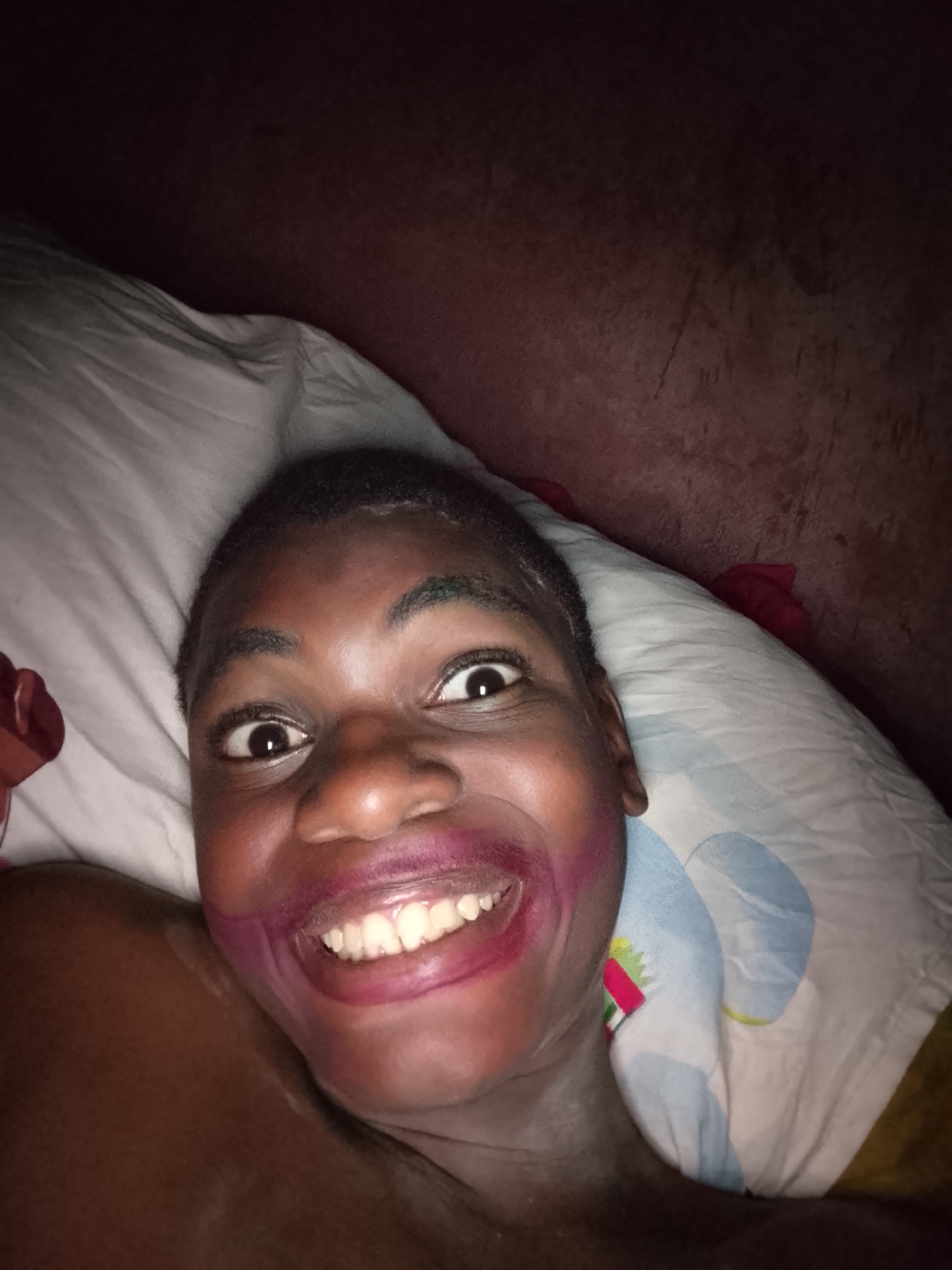A close-up photograph shows a person lying on a pillow. The person has dark skin, short hair, and is smiling broadly, revealing white teeth. There is lipstick smeared around the mouth. The background consists of the pillow, which has a white and blue pattern, and a dark, indistinct surface. The location is Unknown, Unknown.