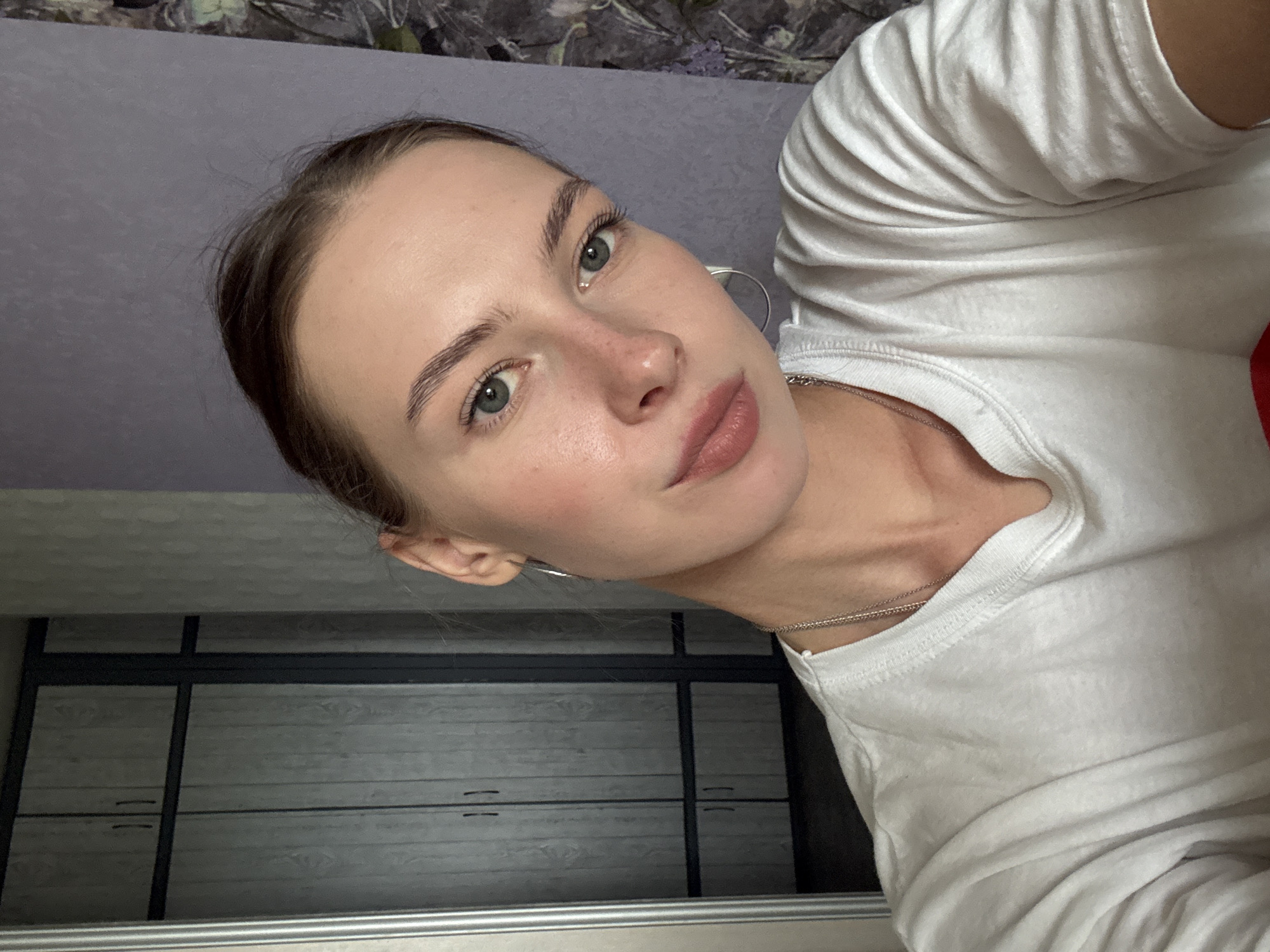 A young woman with light skin, green eyes, and light brown hair tied back is looking directly at the camera in a selfie. She is wearing a white t-shirt and a silver chain necklace with a pendant. She has on hoop earrings. Her makeup appears natural, with emphasis on her eyebrows and lips. The background suggests an indoor setting, possibly a home, with a closet or wardrobe visible on the left and a patterned wallpaper on the right. The lighting is soft, creating a casual atmosphere. There are no specific indicators of the city of Perm, Russia within the image itself.