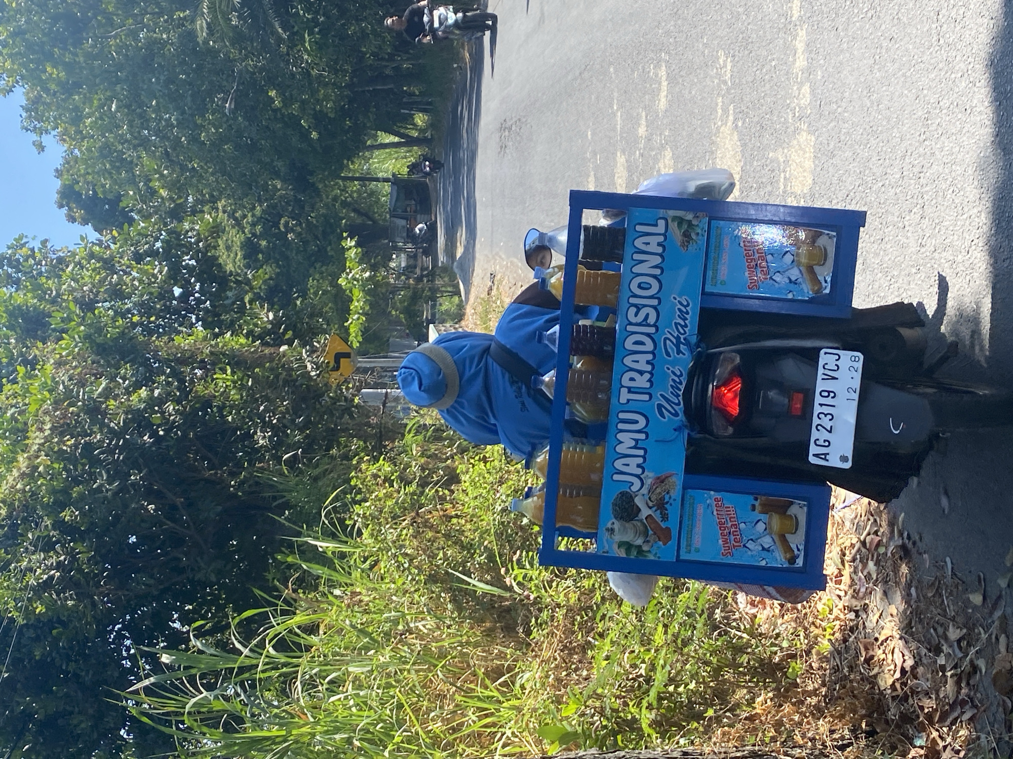 A person in a blue head covering and blue clothing is riding a motorcycle on a paved road. The motorcycle has a blue cart attached to the back, which is filled with bottles of various colored liquids. The cart has a sign on it that reads "JAMU TRADISIONAL Umi Fani". There are also images on the cart depicting ingredients and drinks. The road is lined with lush green trees and foliage. The sky is clear and blue. In the distance, other motorcycles and a small building can be seen. The license plate on the motorcycle reads "AG 2319 VCJ". The scene appears to be in a rural or suburban area, possibly within the city of Kediri, Indonesia, given the Indonesian text on the cart and license plate format.