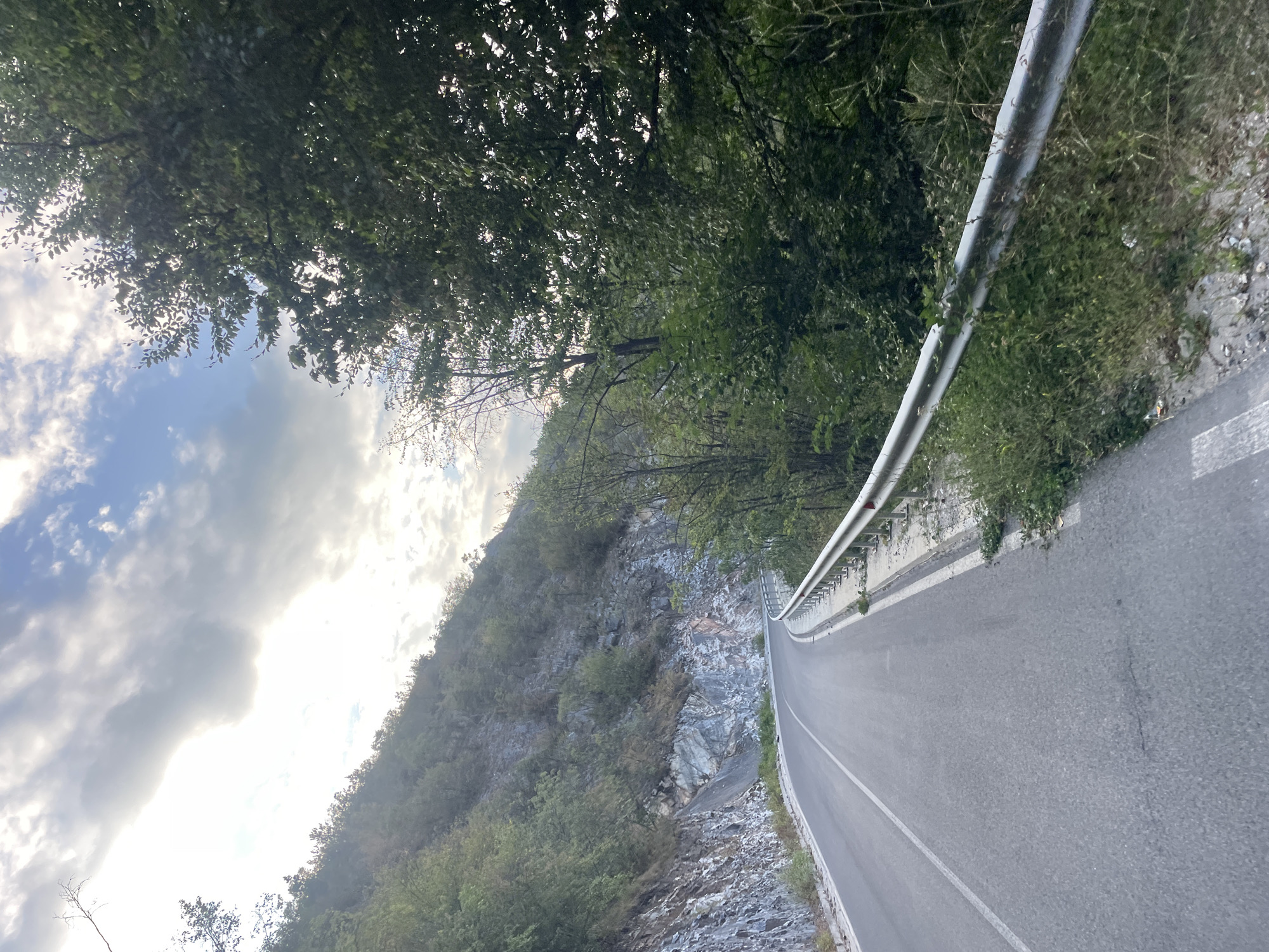 The image shows a winding asphalt road cutting through a mountainous landscape near the city of Yugovo, Bulgaria. The road is bordered by a metal guardrail on the right and a rocky, tree-covered hillside on the left. The sky above is partly cloudy, with sunlight breaking through in patches. Dense green trees and foliage flank the road on the right side, obscuring the background. The road surface appears worn in places, with some debris scattered along the shoulder. The overall impression is one of a scenic, somewhat remote natural setting.