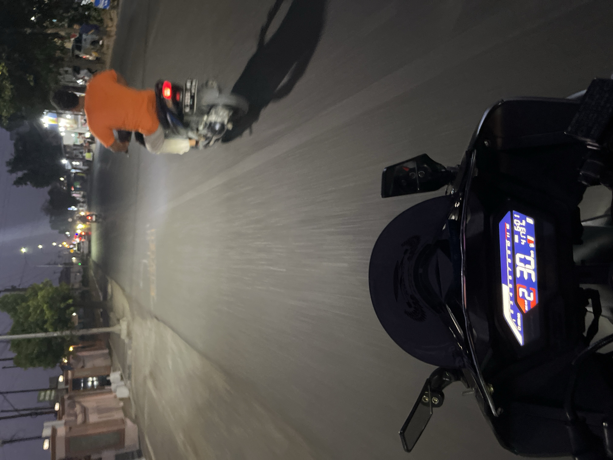The image is a first-person view from a motorcycle at night on a paved road in Kalinyamatan, Indonesia. The foreground shows the motorcycle's digital dashboard, displaying the time as 2:30 AM and other unreadable indicators. A dark windshield is visible above the dashboard, and a side mirror is mounted on the left. In the middle ground, another motorcycle is visible ahead, ridden by a person wearing an orange t-shirt and light-colored pants. This motorcycle has a license plate, though the characters are not clearly discernible. The background depicts a street lined with buildings and trees, illuminated by streetlights and lights from the buildings. The overall scene suggests movement at a moderate speed on a public road during the early morning hours.