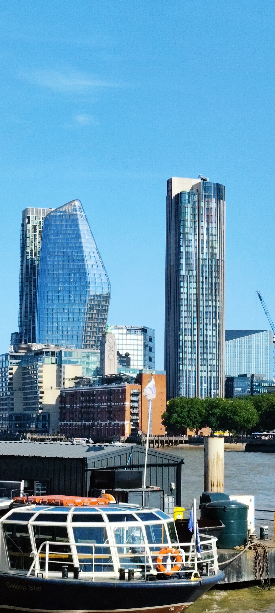 This is a vertical, eye-level, medium-wide shot of the River Thames in London, United Kingdom, on a clear, sunny day. In the foreground, the bow of a dark-hulled boat named "Golden Star" is docked. The boat has a glass-enclosed cabin with orange emergency lights and life preservers visible. Behind the boat is a grey, corrugated metal structure, possibly a warehouse or dock building. A light-colored cylindrical bollard stands next to a dark green water tank on a concrete dock.
In the midground, the London skyline is dominated by modern glass skyscrapers. The most prominent buildings include a tall, rectangular skyscraper with a light-colored cap and a uniquely shaped, blue-glass skyscraper with a curved facade. Several other office buildings with various architectural styles are also visible. A white flag with a blue logo flies from a pole in front of a brick building. Trees line the riverbank behind the buildings.
In the background, a blue construction crane is visible, suggesting ongoing development in the area. The sky is a clear, bright blue with a few wispy clouds. The river is a muted blue-green color, reflecting the sky.