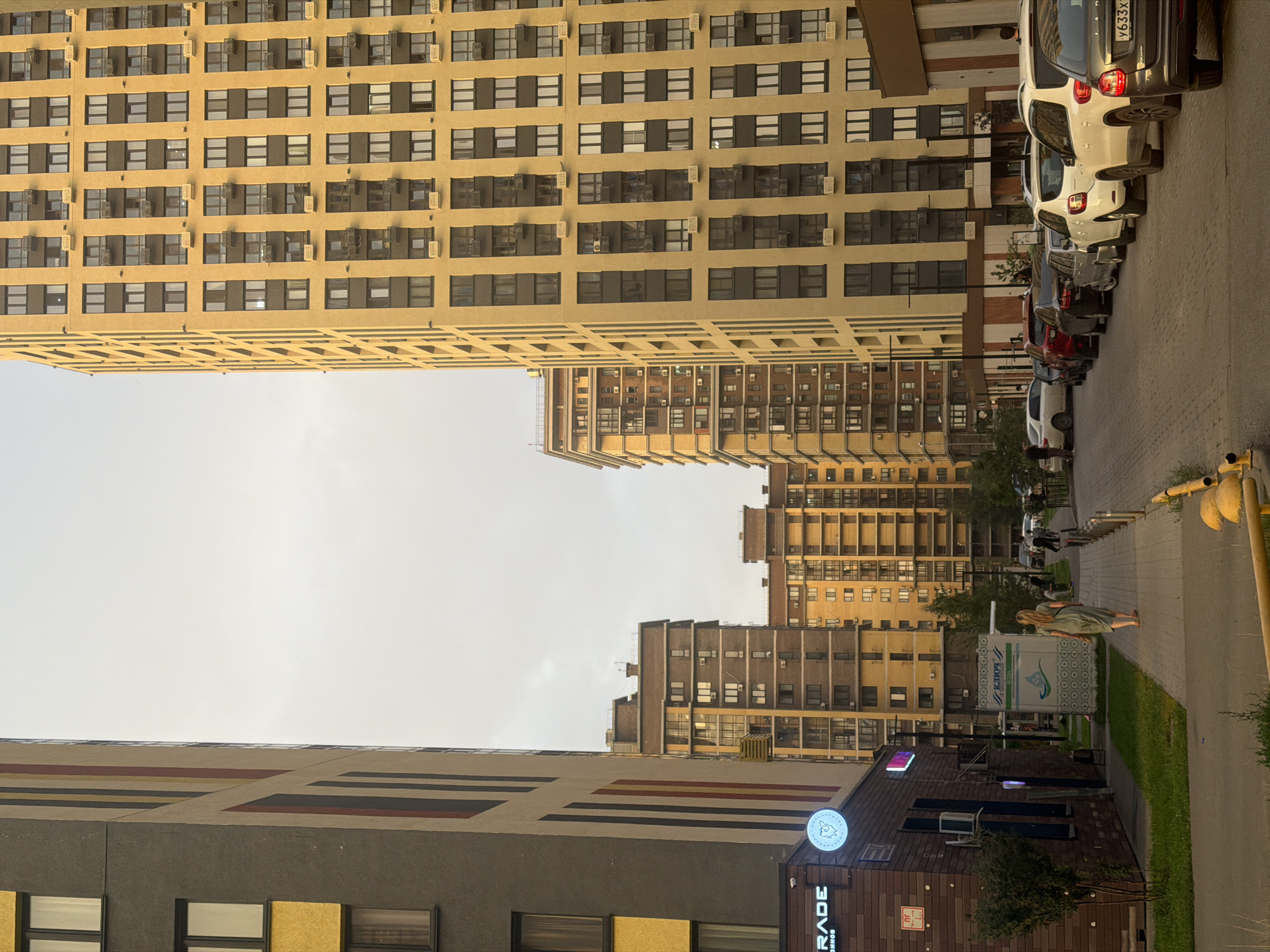 The image depicts a residential courtyard in Kazan, Russia, bathed in the warm, diffused light of what appears to be late afternoon or early evening. Tall, modern apartment buildings dominate the scene, their facades a mix of muted yellow and grey tones, punctuated by numerous windows and air conditioning units. The building on the far right is a prominent, light yellow structure with a grid-like pattern of dark-framed windows. To its left, a similar, though slightly shorter, yellow building is visible, followed by a row of brick-colored buildings, and further to the left, a grey building with bold vertical red and yellow stripes.
A paved walkway runs diagonally through the courtyard, bordered by a strip of green grass on the left and a line of parked cars on the right. Several vehicles are parked along the curb, including a white SUV prominently featured in the foreground on the right, with a visible license plate reading "Y633X". A lone woman, wearing a long, light green dress, walks away from the camera down the paved path. She is barefoot, and her hair is tied back. In the background, other pedestrians can be seen walking, and additional cars are parked throughout the courtyard.
On the left side of the image, adjacent to the grey building, is a low-rise commercial space with a dark brick facade. A lit sign with white text reads "GRADE", and a circular illuminated sign with a stylized image is also present. A small, leafy bush grows in front of this building. Further down the walkway, a stand with the text "КЛЮЧ" and an image of a water droplet is visible, suggesting it is a water vending machine. The sky above is overcast and a pale grey. The overall impression is one of urban residential life with a sense of calm, the soft light and absence of high-activity contributing to this atmosphere.