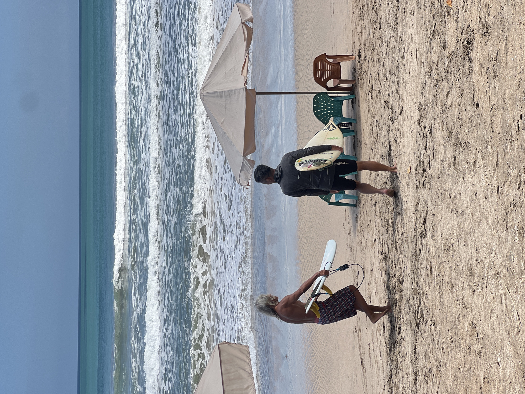In Kuta, Indonesia, two men stand on a sandy beach holding surfboards. The man on the left, with long blonde hair, wears patterned shorts and walks towards the ocean. The man on the right, facing away from the camera, wears a dark long-sleeved shirt and black shorts, holding a surfboard. In the background, the turquoise ocean has gentle waves breaking. Two beach umbrellas, one partially visible on the left and another larger one in the center, provide shade, with several plastic chairs situated beneath the larger umbrella. The sand is damp near the water's edge and drier further up the beach, with footprints visible. The sky is clear and blue.
