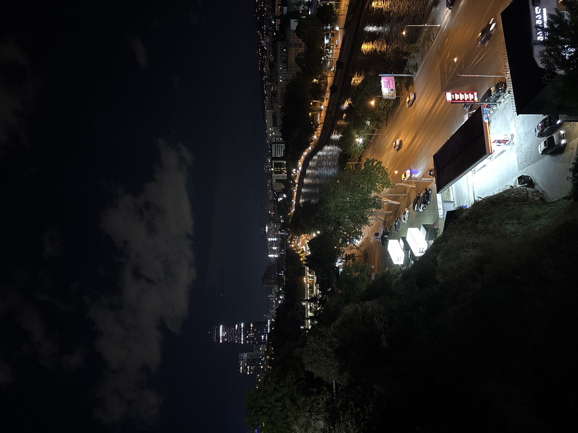 An aerial nighttime view of a city showcases a wide river with reflections of streetlights on its surface. A multi-lane road runs alongside the river, with cars driving on it. On the left side of the road, a gas station with illuminated pumps and a large canopy is visible. Across the river, the city skyline features buildings lit up at night, with a prominent tall skyscraper dominating the view. Trees line the riverbanks and the embankment on the left, obscuring some of the lower areas. The sky above is dark with scattered clouds.