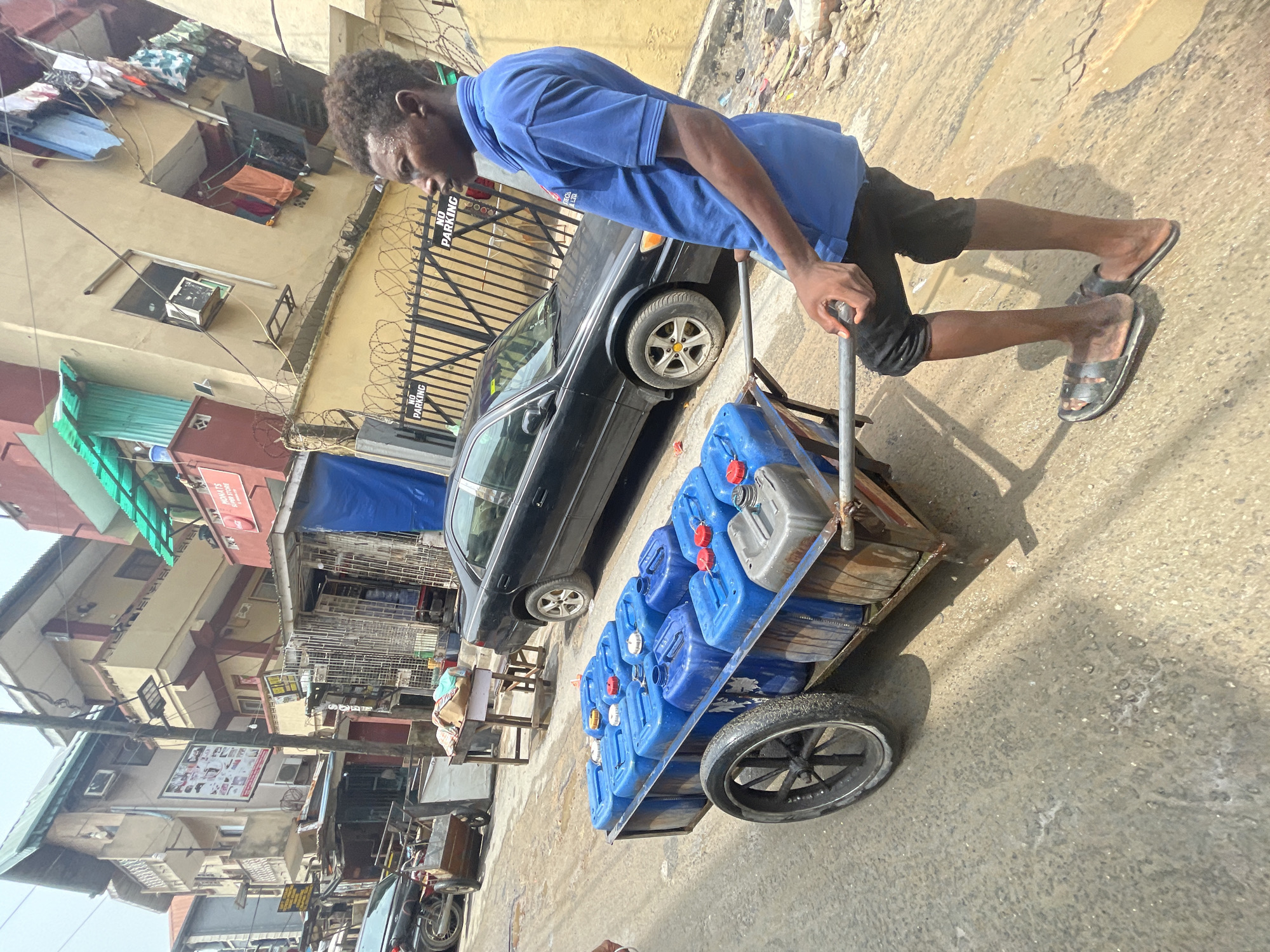 A man in a blue t-shirt and black shorts is pulling a cart filled with blue plastic water containers along a paved street. He is wearing sandals. The cart has two large, spoked wheels. A black sedan is parked nearby. The backdrop includes buildings with apartments and commercial establishments. There is barbed wire atop a fence near the parked car. The location is suggestive of Lagos, Nigeria, indicated by the architectural style and street scene.