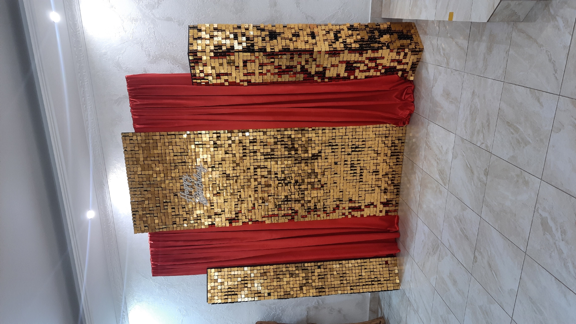 The image shows a party backdrop with a sequined wall and red curtains. The backdrop is set up against a textured white wall and is placed on a tiled floor. There is a silver "Happy Birthday" sign on the sequined wall. The backdrop consists of four vertical sequined panels of varying heights, with red curtains draped between them. Two of the sequined panels are rectangular, while two are square. The sequined panels are made of small, square, gold and black sequins that reflect light. The red curtains are made of a smooth, satin-like material and are gathered at the bottom. The backdrop appears to be set up indoors, likely for a celebration. The specific location in the city of Жодзішкаўскі сельскі Савет, Belarus, cannot be determined from the image alone. There are no people visible in the image.