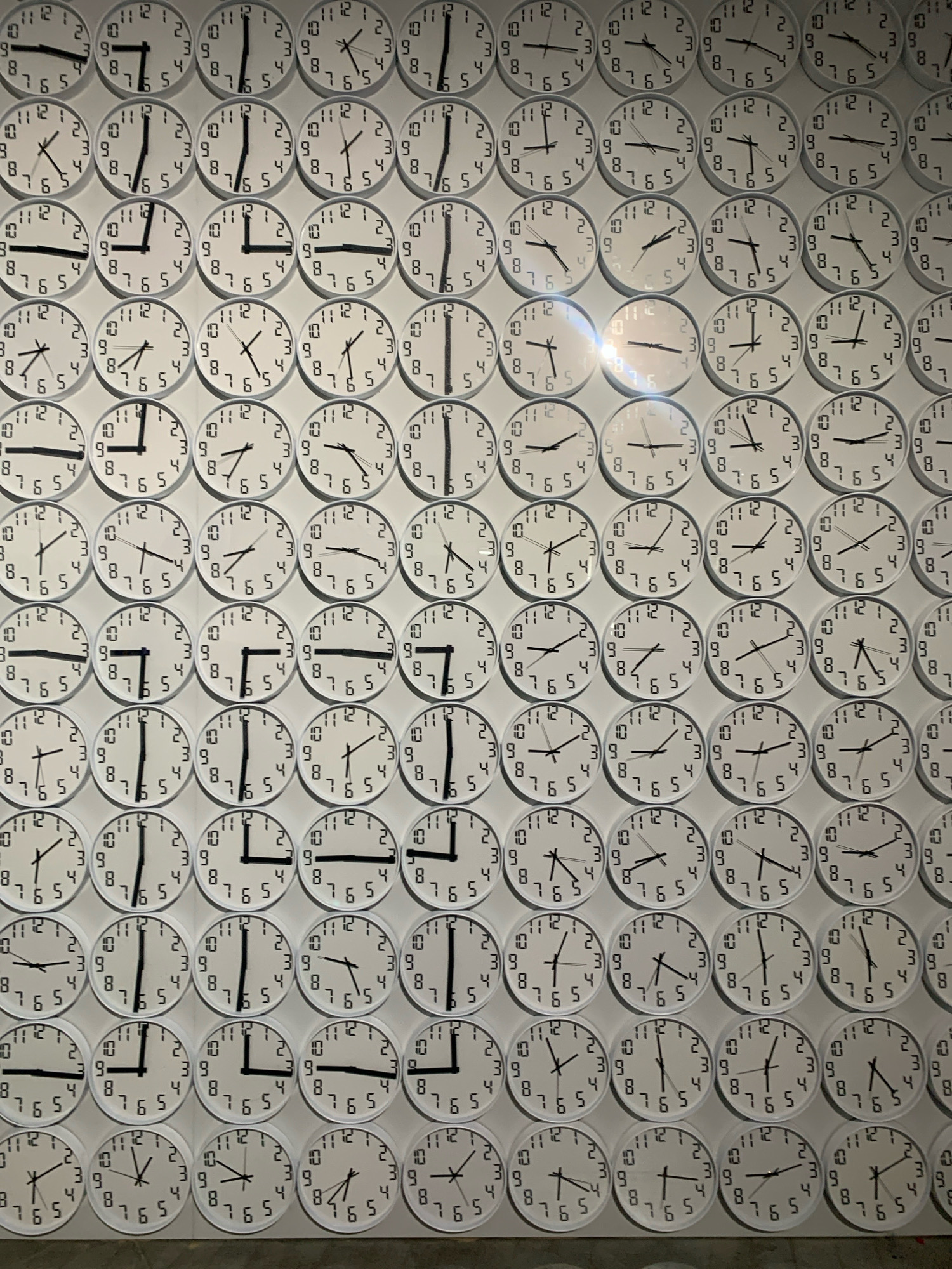 The image shows a wall covered with a large number of analog clocks. The clocks are arranged in rows and columns, and each clock displays a different time. The clocks are all white with black hands and numbers. The background is a light gray wall. There are no people visible in the image. The image does not contain any information that would allow for the determination of its location in Kyiv, Ukraine, or any other city.