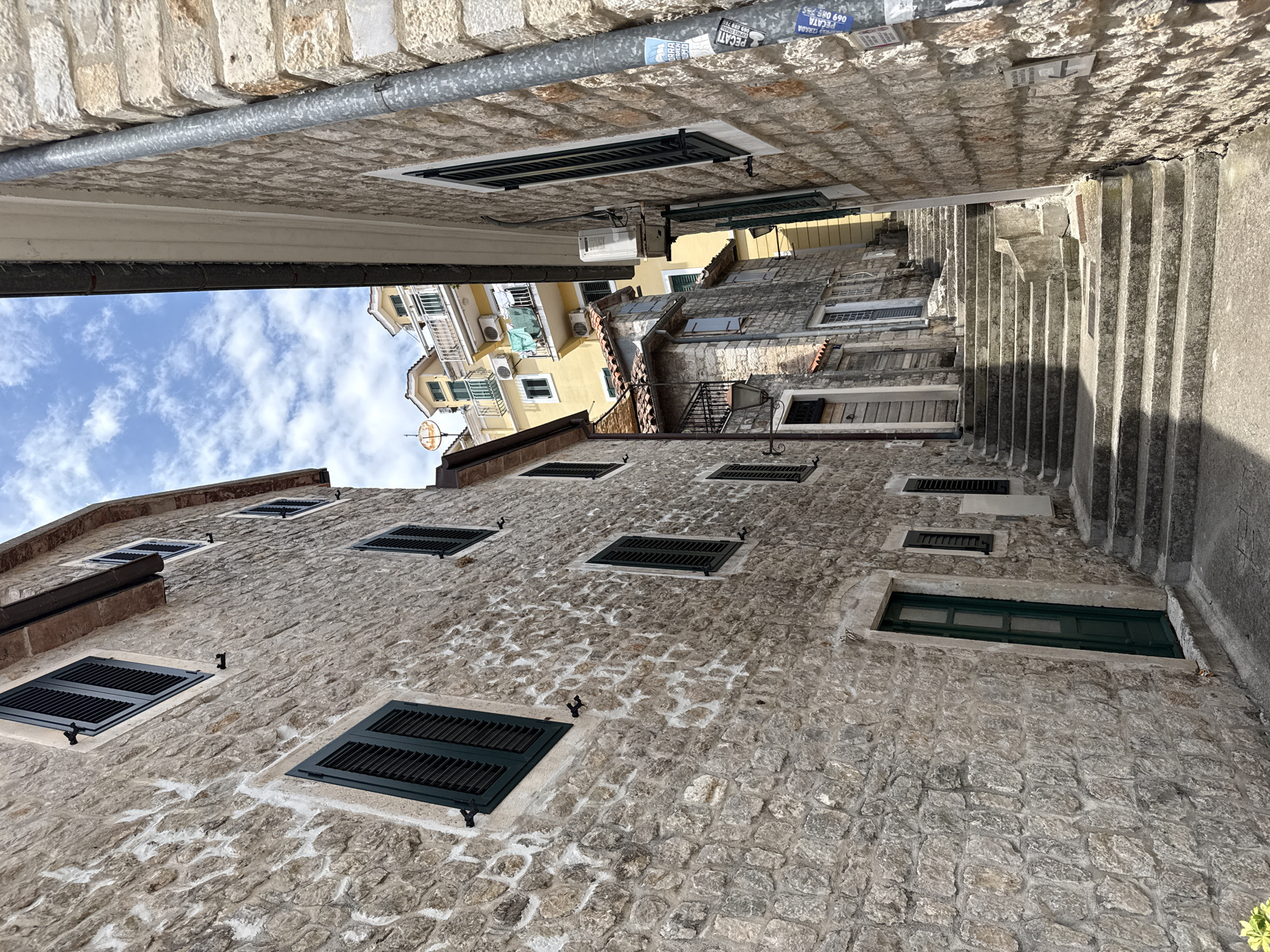 A narrow stone alleyway in Herceg Novi, Montenegro, is depicted from a low angle, looking upwards. The walls of the buildings are constructed from rough, light-colored stone with visible mortar. Dark green shutters cover most of the windows, which are visible on both sides of the alley. The alleyway itself is a set of stone stairs ascending towards a brighter area at the top, suggesting it leads to a higher part of the city. The sky above is a vibrant blue with scattered white clouds. The scene is quiet and appears to be uninhabited. The architecture suggests a historic Mediterranean town.