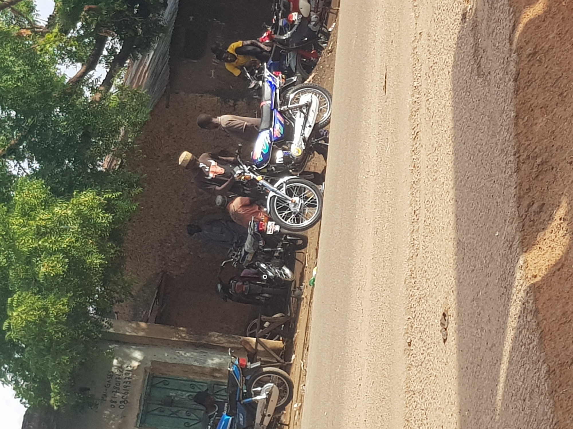 A group of people are gathered around motorcycles in front of a building in Kufena, Nigeria. A blue motorcycle is parked on the left, and several other motorcycles are visible in the background. Three men are standing near the motorcycles, and one is seated on a motorcycle. A tree with green leaves shades the area. The building appears to be made of mud and has a green door. The ground is paved, and a dirt road is visible in the foreground. The image is taken from a slightly elevated angle, suggesting the photographer is standing on the road.