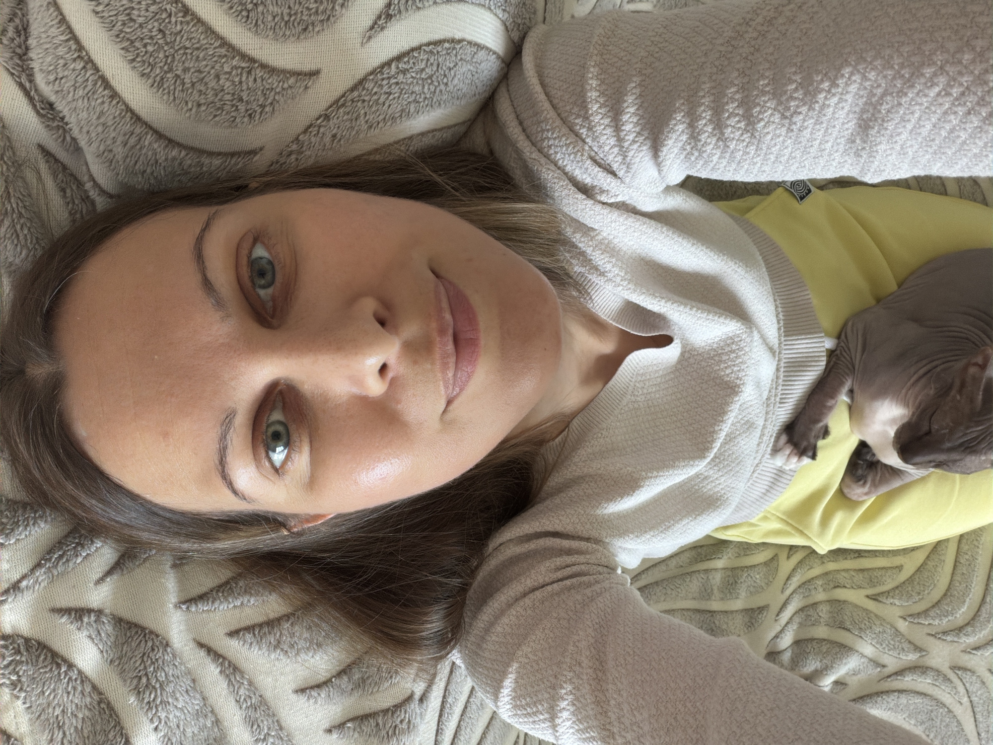 In Izhevsk, Russia, a woman with light brown hair is lying down on a textured blanket. She is wearing a light-colored sweater and yellow bottoms. A Sphynx cat is resting on her lap. The woman is looking at the camera with a slight smile.