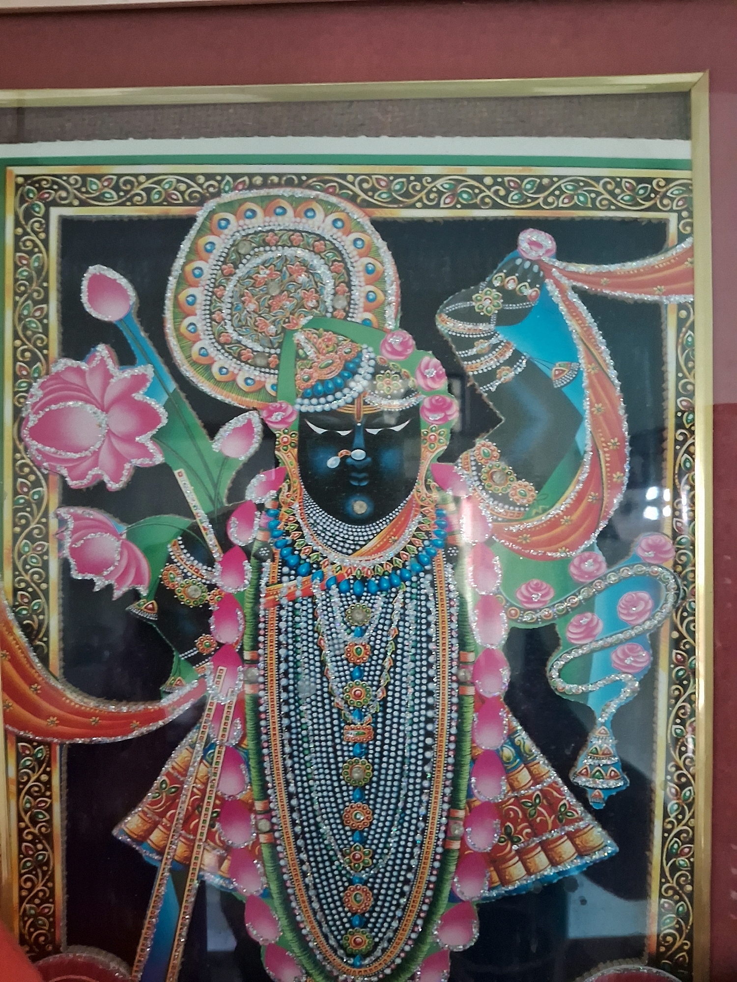 The image shows a framed painting of a deity, likely from Hindu mythology, with dark skin, adorned in ornate jewelry and vibrant clothing. The deity is depicted centrally, with elaborate floral motifs and decorative patterns surrounding them. The painting is highly detailed with what appears to be small, sparkling embellishments. The framing is gold-colored with intricate borders. The background of the painting is black. The photograph is taken from a slightly downward angle, showing a portion of a dark reddish frame at the top. The painting is displayed indoors. This painting is a religious artwork and could be found in temples, homes, or art galleries in Udaipur, India.