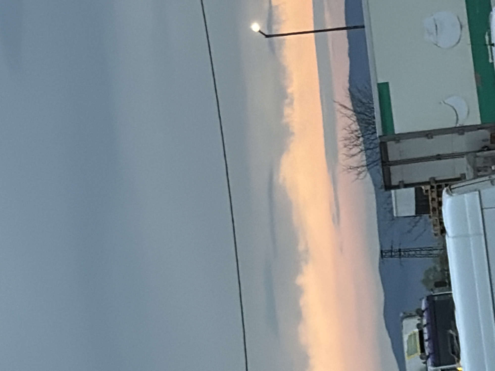 This image captures an outdoor scene at either sunrise or sunset, featuring a vibrant sky above an industrial or commercial landscape. The sky on the left half of the frame is a muted blue-grey, transitioning towards the center and right into soft, peachy-orange and pink hues, indicative of low sun angle. Diffused clouds are visible within these warmer tones.
A thin, dark power line or cable stretches diagonally across the upper portion of the sky. Near the top center, a street light with a brightly illuminated bulb extends into the frame. Along the horizon, a dark, undulating silhouette of distant mountains or hills separates the sky from the foreground elements.
On the right side of the image, man-made structures are visible. A light-colored building, possibly a warehouse, occupies the upper-right, featuring green trim and several white circular or curved shapes on its surface. Below this, a network of dark, bare tree branches is silhouetted against the colorful sky. In the mid-ground, a tall, dark metal lattice tower, likely a power line pylon, stands against the distant mountains.
Vehicles are present on the far right; the side and rear of a large white truck or van are partially cropped into the foreground. Other indistinct trucks or trailers are parked behind it. The scene presents a blend of natural beauty and industrial utility, with no visible text.