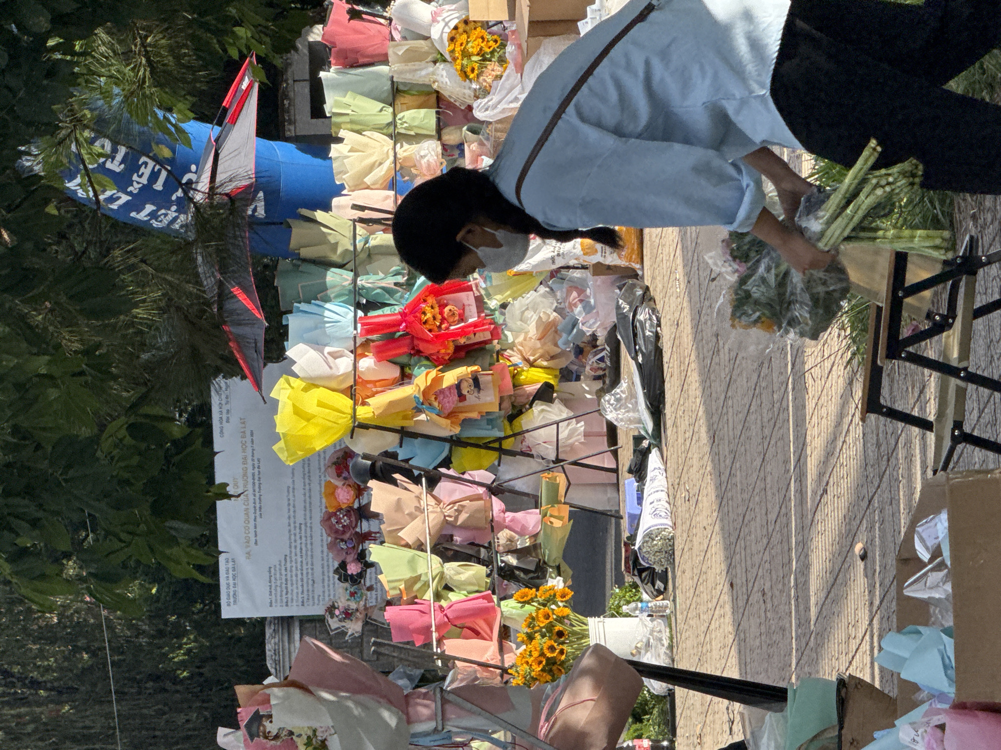 In the city of Lâm Viên - Đà Lạt, Vietnam, a street vendor is seen arranging a bouquet of flowers on a sunny day. The vendor, a young woman with her hair in a braid and wearing a light blue shirt and a white face mask, is crouched down and holding the stems of a large bouquet of green leafy flowers, possibly asparagus, in her hands. She is surrounded by various other flower bouquets wrapped in colorful paper, which are displayed on metal racks. In the background, a blue inflatable balloon with text written in Vietnamese can be seen, along with trees and a sign board. The ground is paved with square tiles, and several boxes and plastic bags are scattered around the scene, suggesting a bustling market environment.
