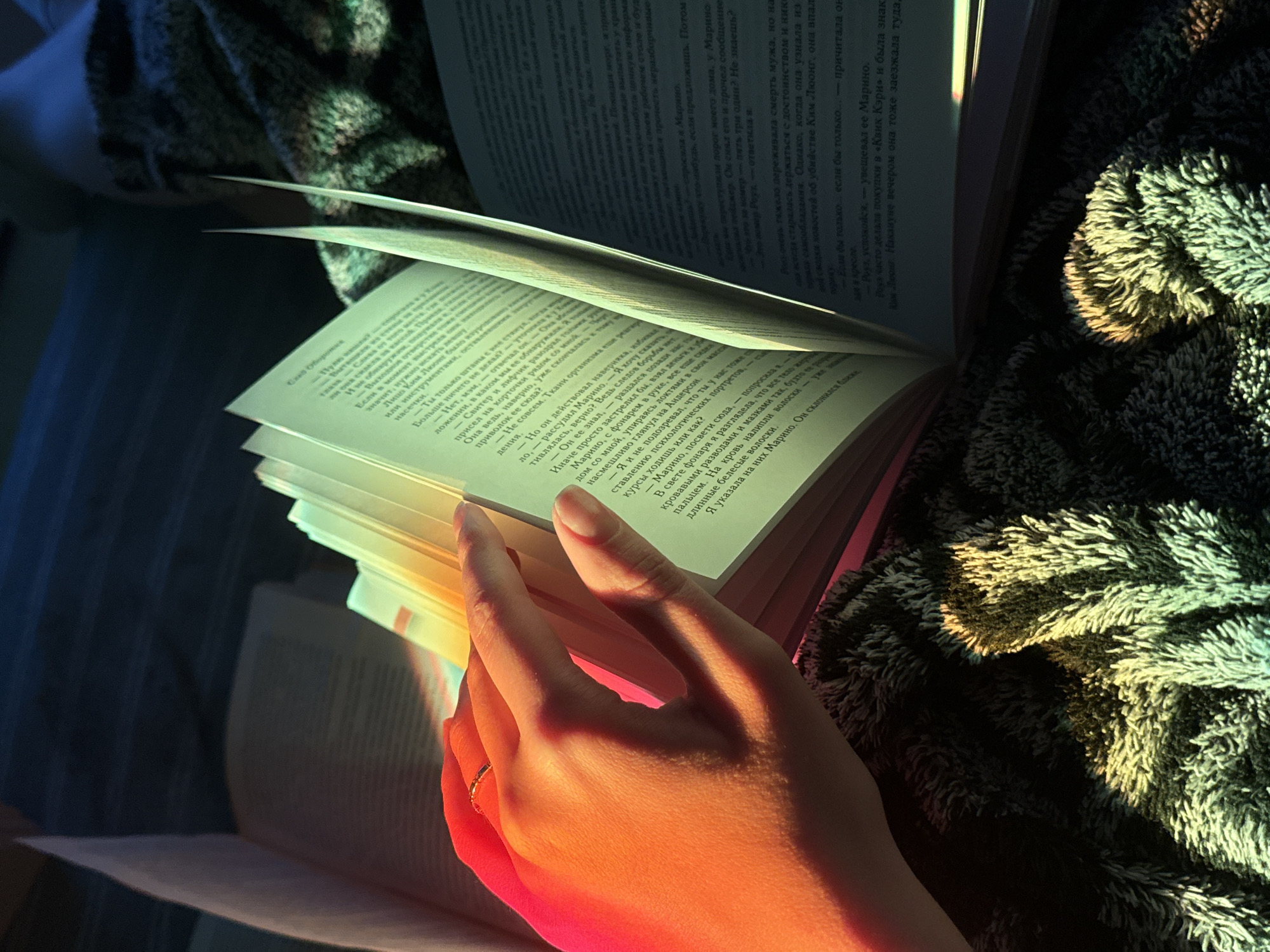A close-up shot captures a hand with a ring, gently turning the pages of an open book. The book appears to be in Russian, with dense text on the pages visible. The scene is bathed in a warm, somewhat surreal, light, with strong orange and pink hues highlighting the hand and pages closest to the viewer. A textured material, possibly a blanket or cushion, can be seen near the book, with a mix of light and shadow adding depth to the image. The background is a mix of blue and shadow, suggesting a dimly lit room or a low-light setting.