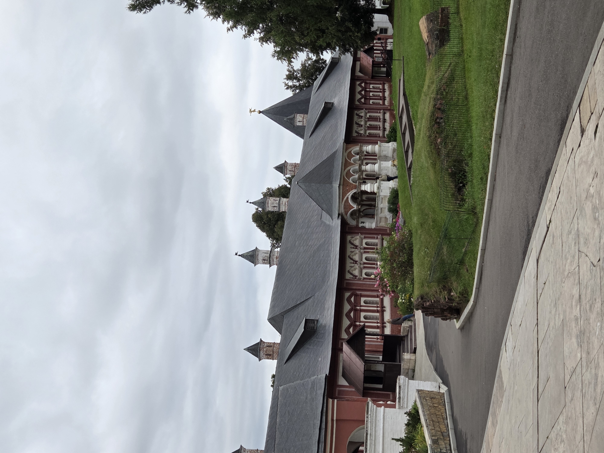 The image shows a view of Savvino-Storozhevsky Monastery in Zvenigorod, Russia. The monastery's red and white architecture features multiple small towers with pointed roofs and a larger, dark-colored, pointed central tower topped with a golden emblem. The building has a long, pitched, dark grey roof with dormer windows. A paved path curves from the foreground towards the monastery entrance, which is marked by a covered porch with stairs. To the right of the path is a grassy area with a low stone wall and a dark metal fence. Several trees, including a large evergreen, are visible behind the monastery and to the right. The sky is overcast with grey clouds. A lone person is sitting on the stairs to the right of the entrance.