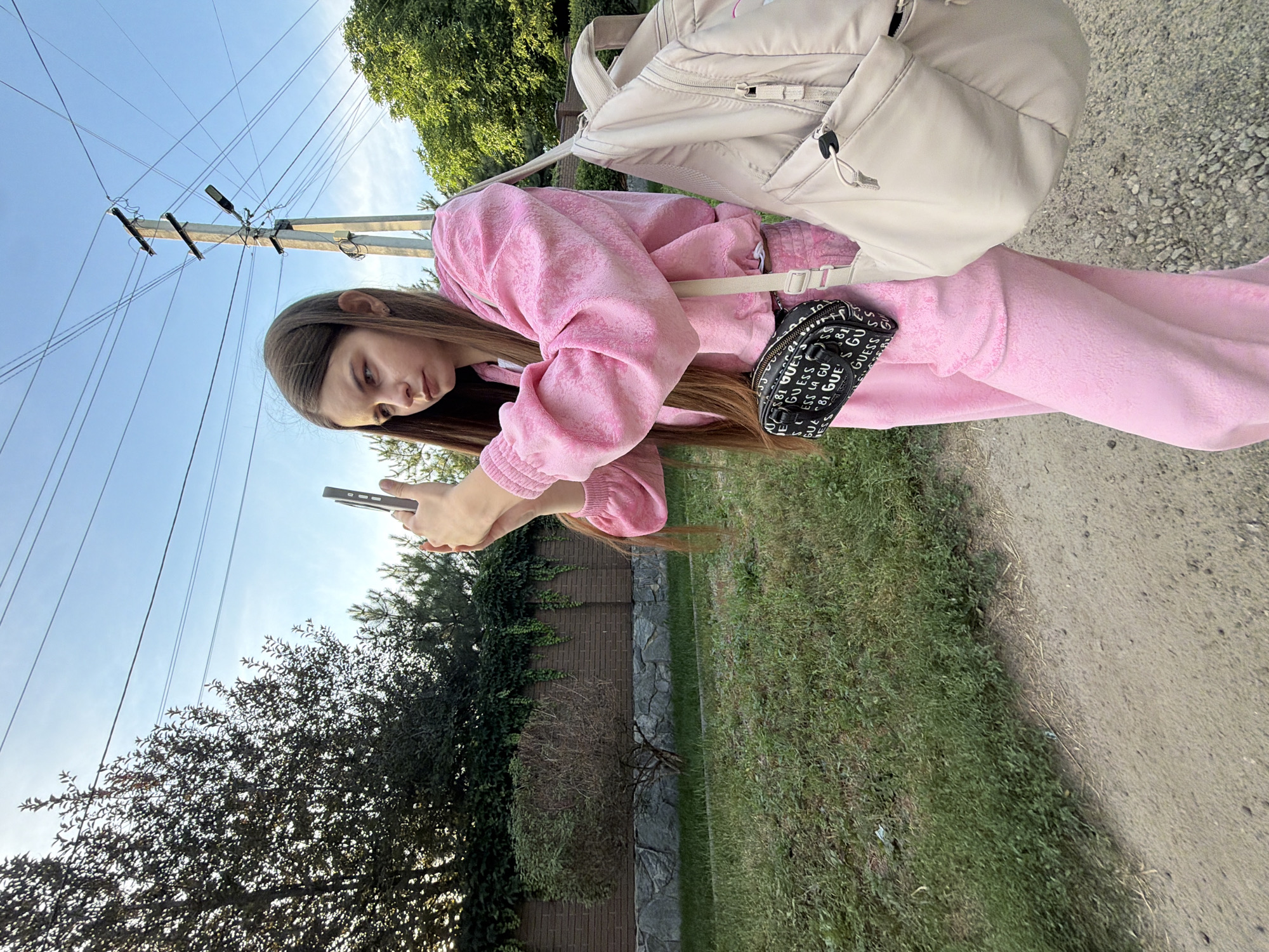 A young woman wearing a pink tracksuit and a light-colored backpack is standing outdoors, holding a smartphone. She is looking down at the phone, seemingly engaged in using it. A black fanny pack with "GUESS" logo pattern is visible around her waist. The scene appears to be a bright, sunny day, suggested by the clear blue sky and the strong sunlight creating shadows. The environment includes a gravel path, green grass, and trees, with a brick and stone fence in the background. Utility poles and wires are also prominent in the sky. The image seems to be taken from a low angle, looking up towards the sky and the woman. The overall impression is one of a casual outdoor moment.