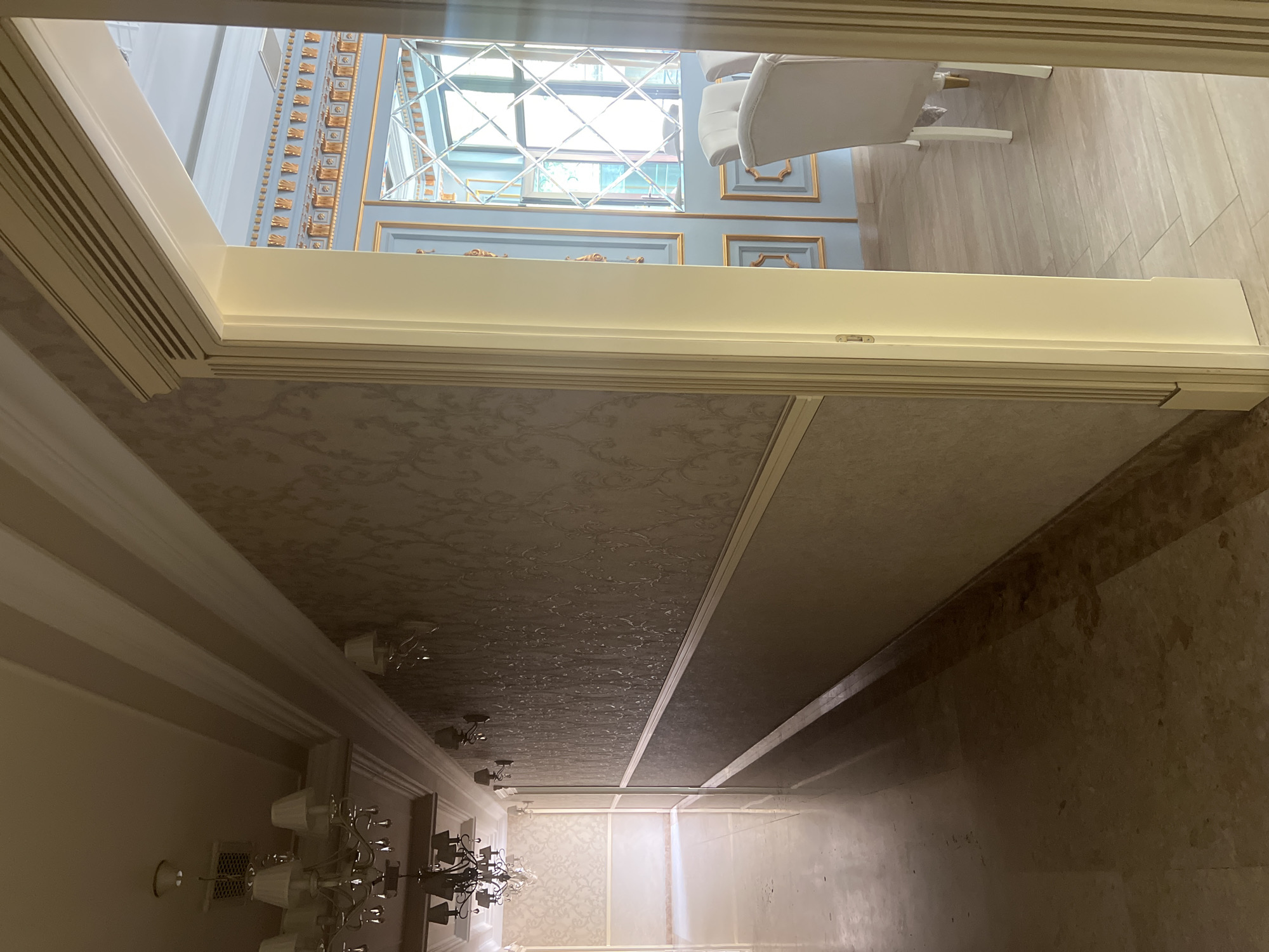The media file captures a luxurious interior scene, looking through a white archway or doorway into an adjacent room. In the foreground room, a patterned ceiling, possibly wallpapered, features an ornate white chandelier with multiple lights and shades.
Beyond the archway, the second room is distinguished by light blue walls adorned with intricate golden decorative molding, creating framed panels. A prominent, large mirror with a geometric, crisscross patterned frame reflects bright light, suggesting the presence of a window or significant light source. A white, plush chair is partially visible, indicating a seating area. The floor in this room is covered with light-colored, possibly beige or cream, rectangular tiles.
The overall aesthetic is one of opulence and classical elegance. No people are visible, and no specific activity is taking place. The bright reflection suggests it is daytime. No text is visible in the image. The setting is an interior space, likely in Yerevan, Armenia.