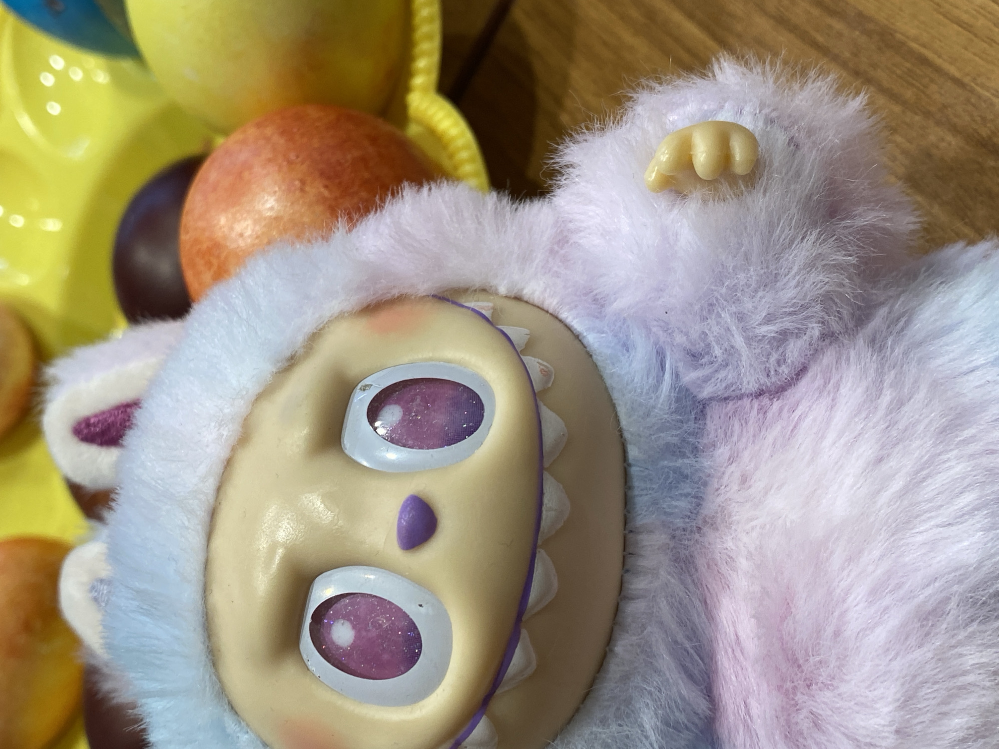 A close-up shot reveals a Furby-like toy with a plastic face and faux fur. The face is light yellow with purple eyes and a triangular nose. The fur is a mix of white, blue, and purple. In the background, there's a yellow plastic tray with colorful balls. The setting seems to be indoors on a wooden surface. There is no evidence to suggest the location is in Votkinsk, Russia.