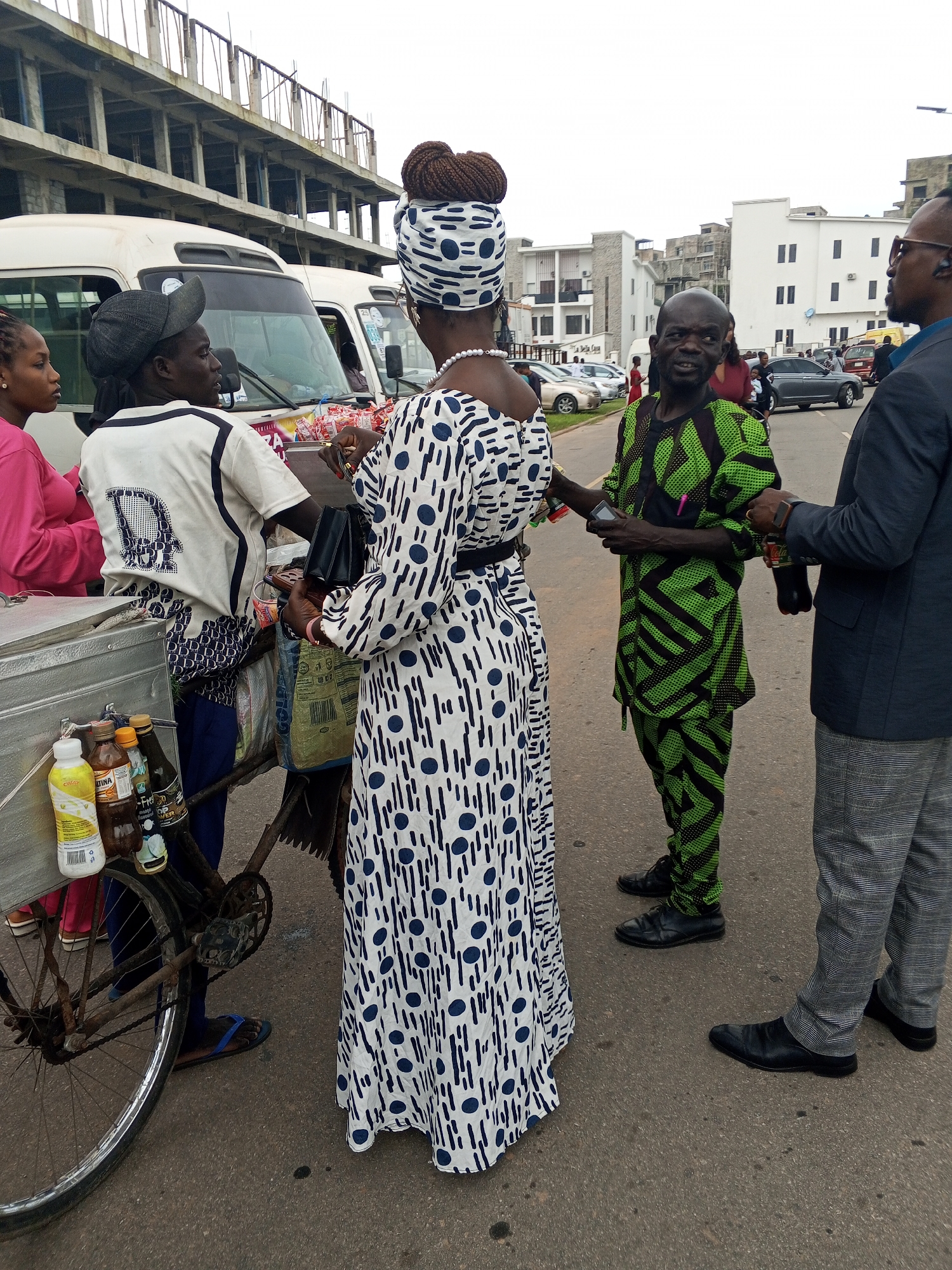 The image depicts a street scene in Guzape, Nigeria, with several individuals gathered around a mobile vending cart. In the foreground, a woman wearing a white dress with blue polka dots and a matching headscarf stands facing away from the camera. Next to her, to her left, is another person in a white shirt with a dark pattern and a dark cap, also looking towards the right. A woman in a pink top is visible on the far left, partially obscured.
The vending cart, a metal box on a bicycle frame, holds several bottles of drinks. Behind these individuals, a man in a vibrant green and black geometric-patterned traditional outfit is speaking with another man. The man in the green outfit is looking directly at the camera, while the other man, in a dark suit jacket and grey checkered pants, is looking to his right. The background shows a street with parked cars, modern buildings, and an unfinished concrete structure. The sky is overcast. The people appear to be engaged in a transaction or conversation.