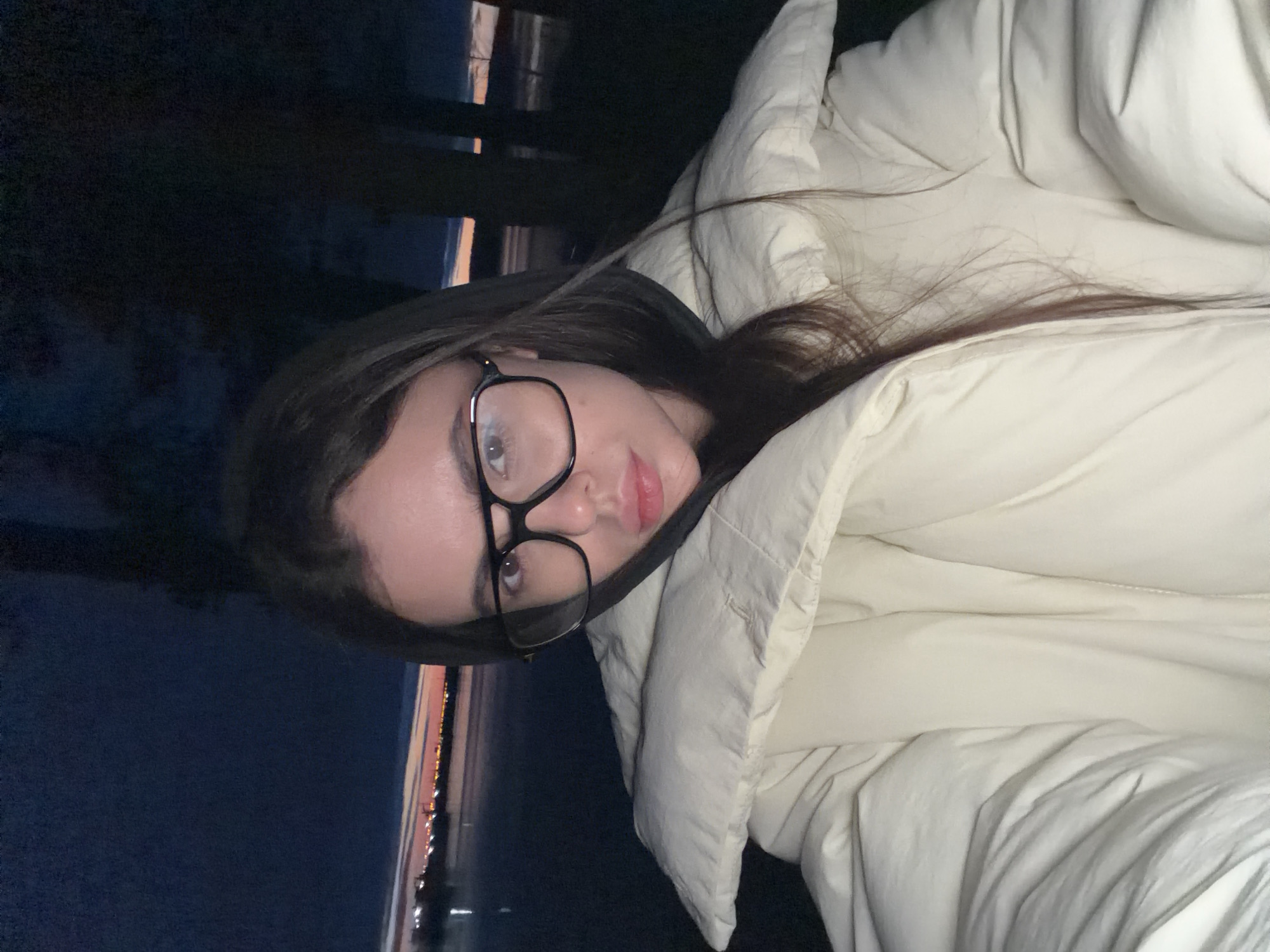 A woman with long brown hair and glasses is depicted in a medium shot wearing a light-colored, puffy jacket. The background shows a nighttime scene with dark blue sky and a faint glow on the horizon, suggesting a possible body of water. Lights, possibly from buildings or streetlights, are visible on the horizon. There is a fence or railing behind the woman. The city of Chaikovsky, Russia, is not discernible from this image.