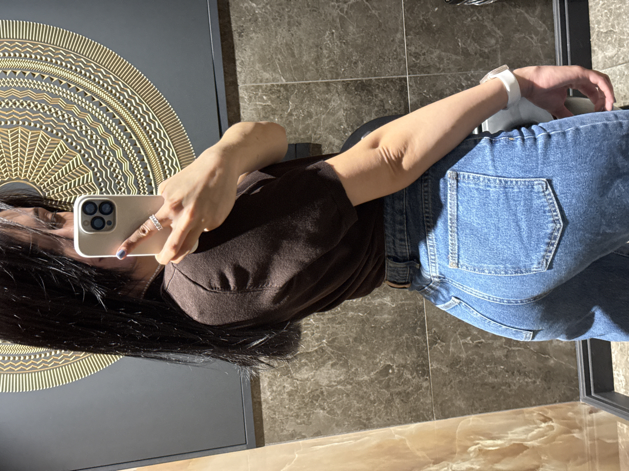 A person is seen in a selfie taken in front of a mirror. The individual is wearing a brown shirt and blue jeans. A white watch is on their wrist. The person is holding a smartphone in their left hand, taking the picture. The background includes a wall with a decorative golden design, a floor with a marbled texture, and a dark-colored wall or furniture piece. The location is unknown; there is no indication that the photo was taken in the city of Votkinsk, Russia.